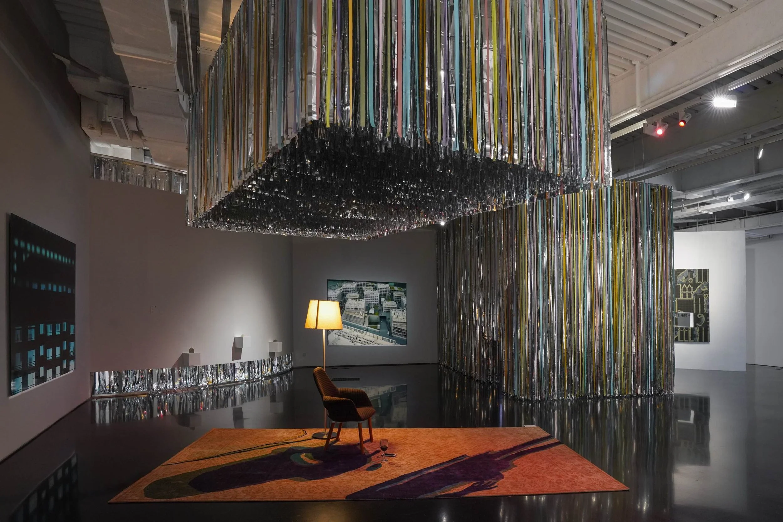 Art installation with hanging colorful ribbons in an art gallery, with a yellow lamp, chairs, and a patterned rug on the floor.
