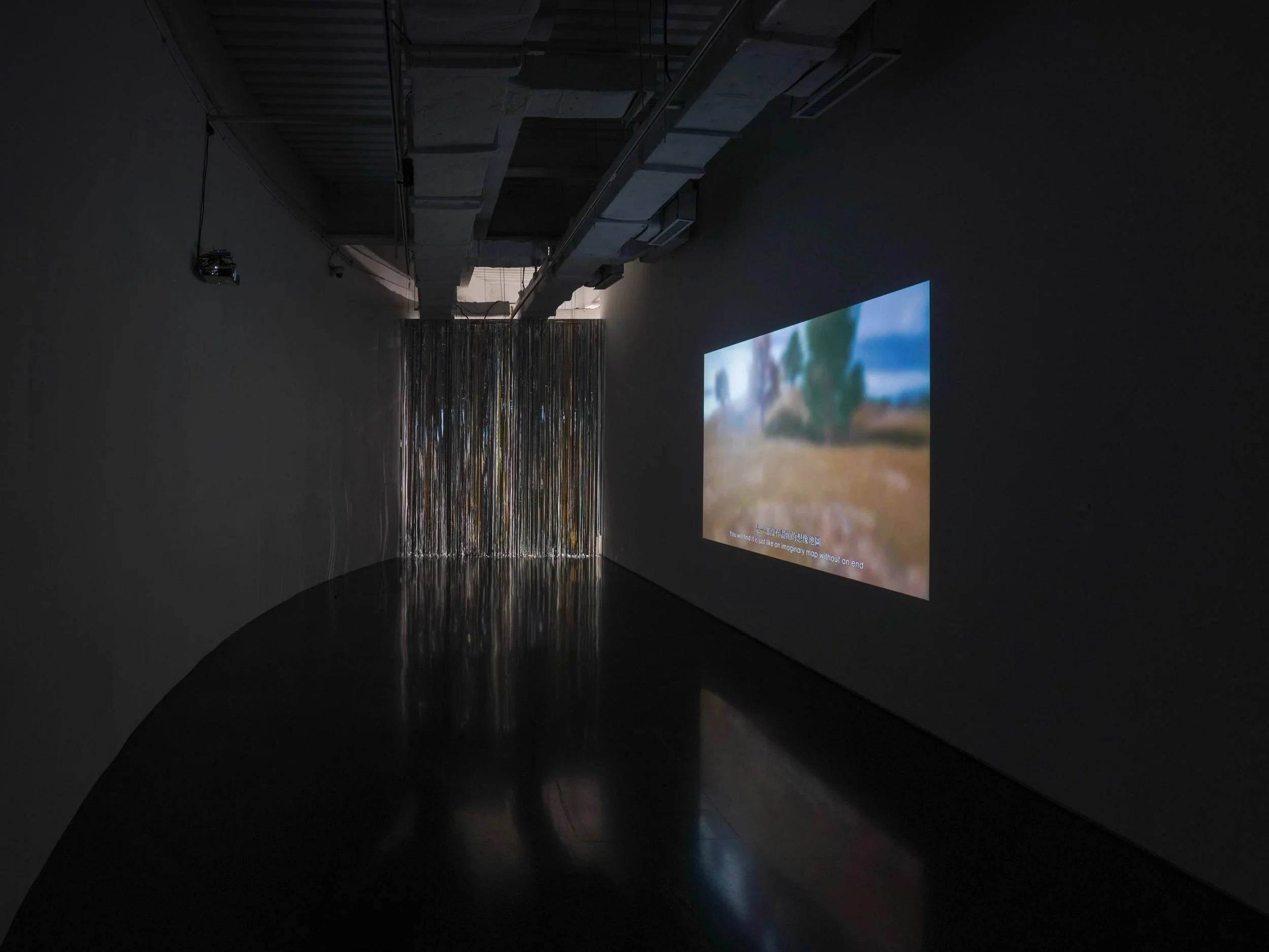 Installation view of Danwei Unfolding (2025) at TANK Shanghai, featuring a large-scale video projection of a landscape-like scene on a dark gallery wall, with Nishiko's suspended multicoloured ribbons visible at the far end of the space.