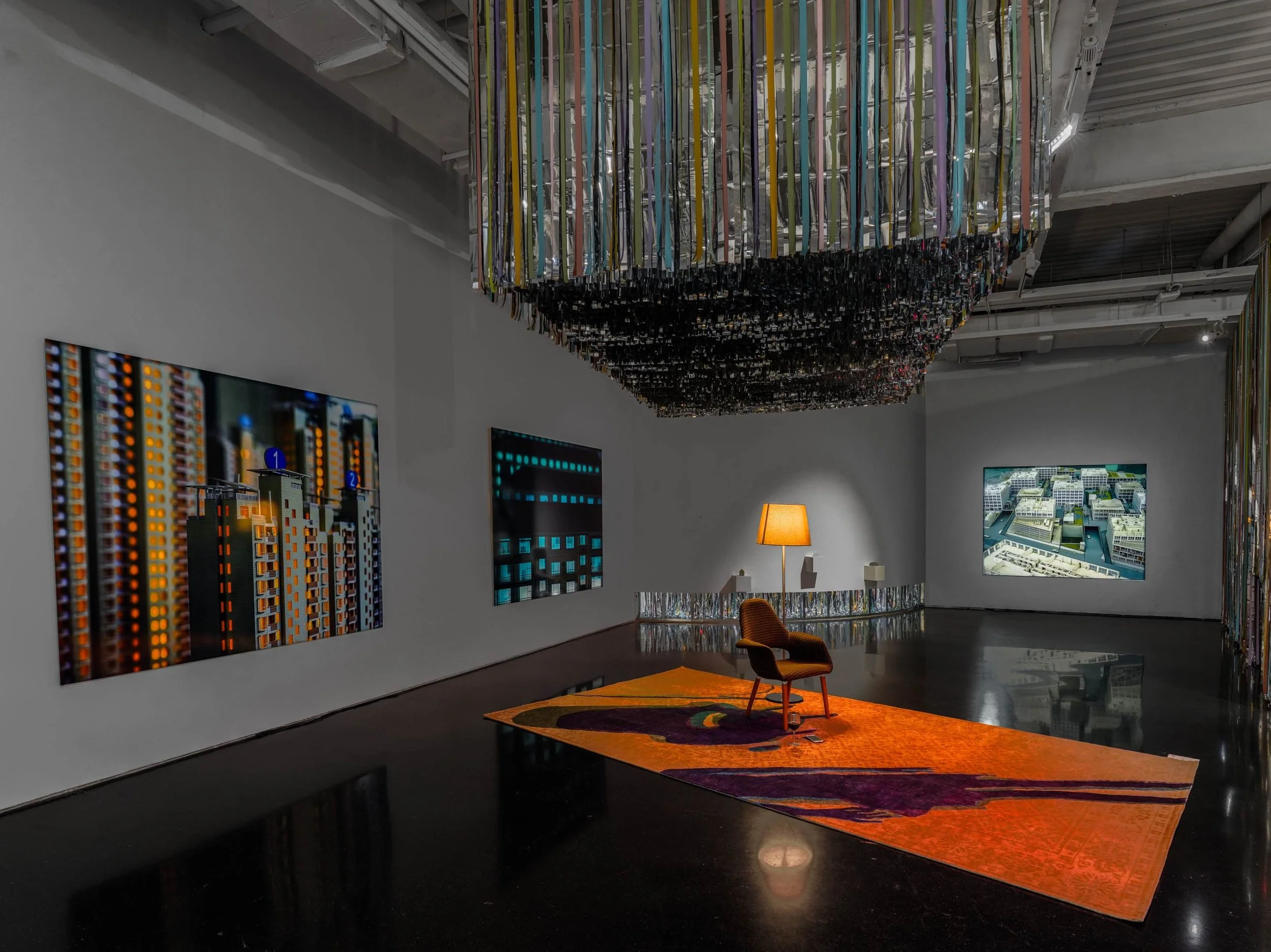 An art gallery with a large installation hanging from the ceiling, colorful row of ribbons or streamers. Walls display digital artwork or photographs, a lamp on a table, and a patterned rug on the floor by artist Xing Danwen.