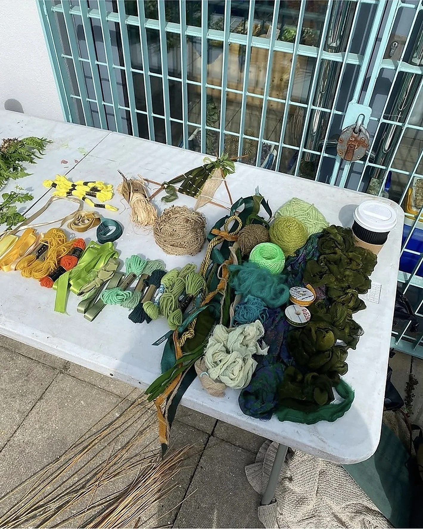 Colourful yarns, textiles, branches and other natural materials arranged on an outdoor table for a collective making workshop led by Hannah Lees and May Hands as part of To Be Here With You (2023) at Southwark Park Galleries, London.