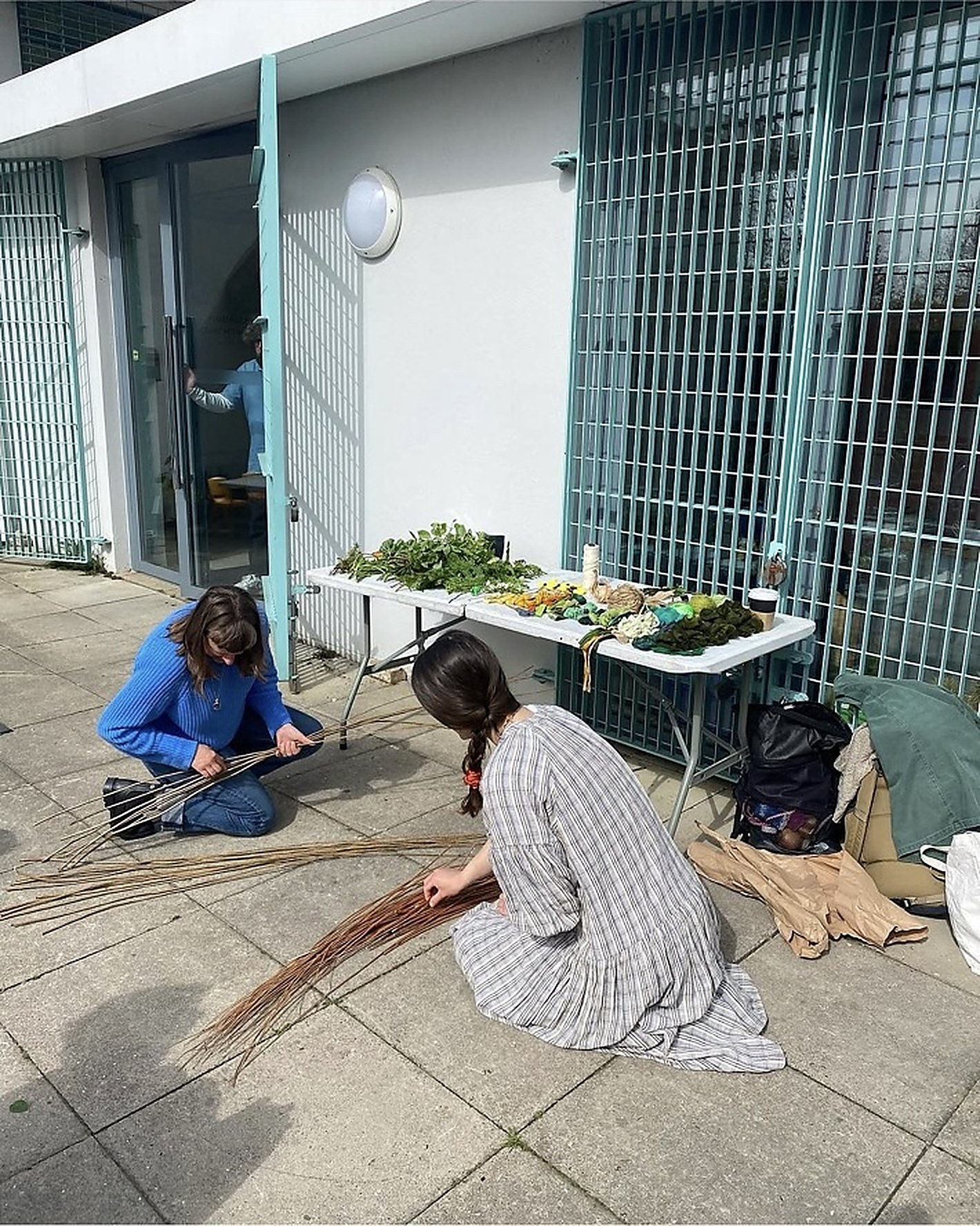 Two participants working with and combining natural and foraged materials during an artist-led workshop by Hannah Lees and May Hands as at Southwark Park Galleries, London, with plants and workshop supplies arranged nearby.