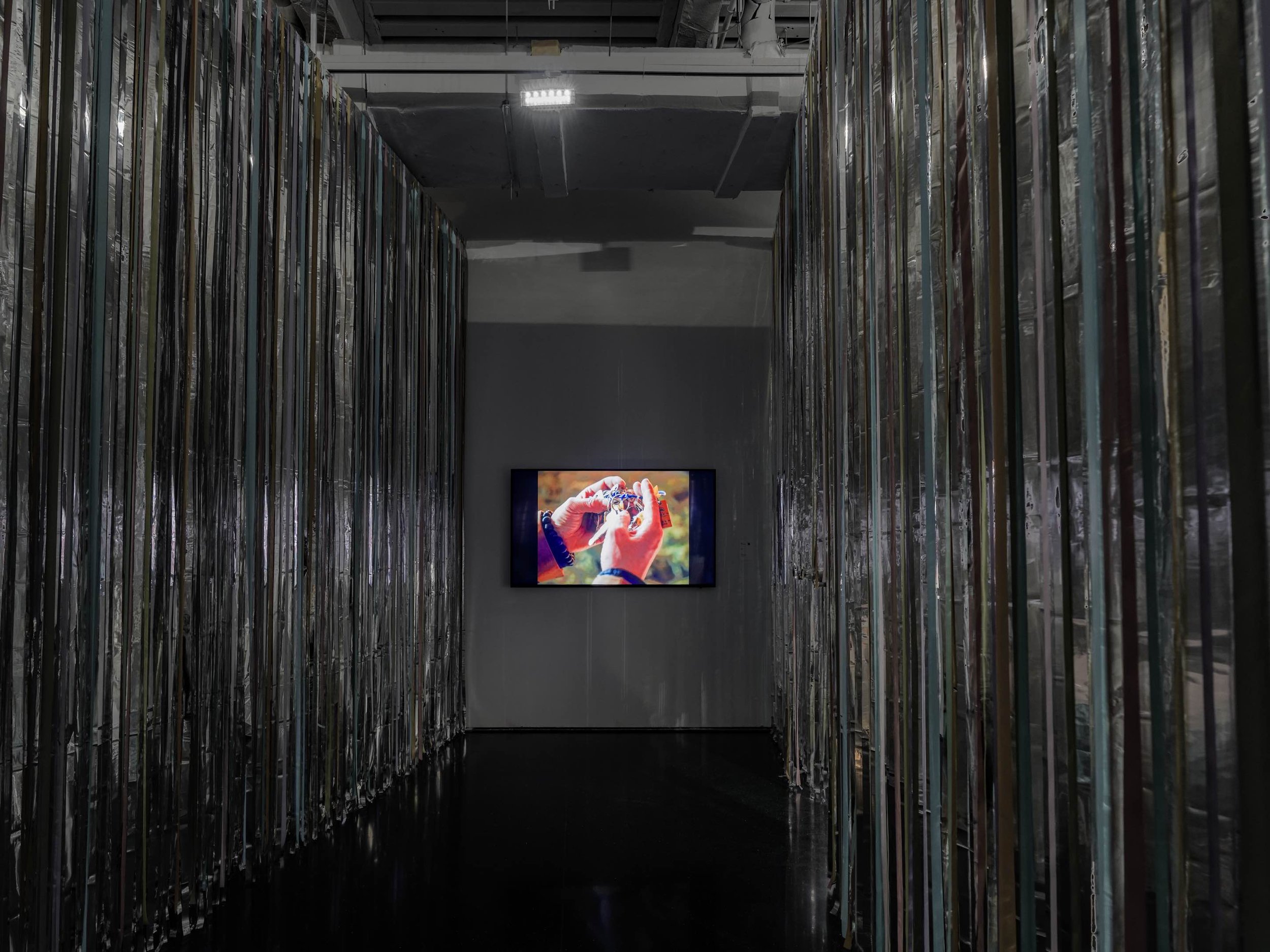Installation view of a video work by Kanthy Peng in Danwei Unfolding (2025) at TANK Shanghai, displayed on a screen at the end of a dark passageway enclosed by Nishiko's suspended multicoloured ribbons.