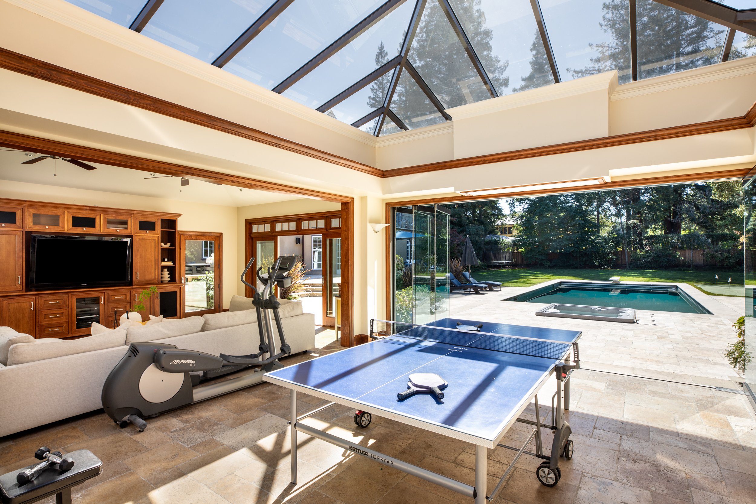 Interior view of a spacious living room with large windows and a glass ceiling, showcasing an outdoor pool and a backyard with trees.