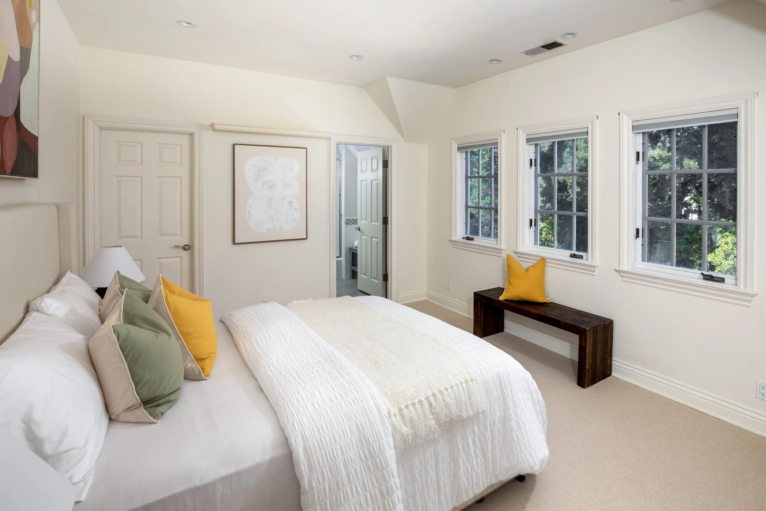 A bedroom with a large bed, white bedding, and colorful pillows, three windows with trees outside, a wooden bench with a yellow pillow, and some wall art.