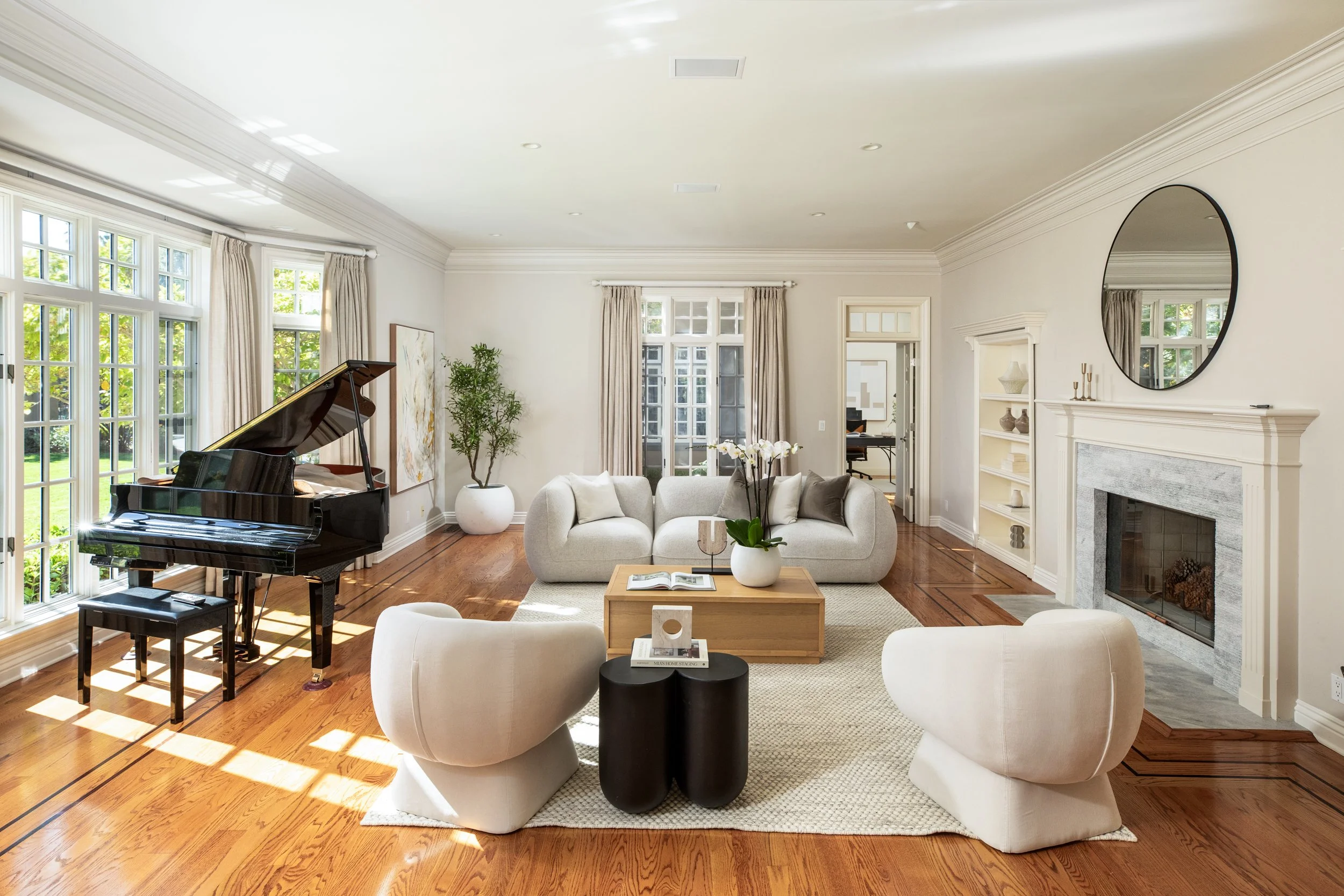 A spacious living room with large windows, hardwood floors, and white walls. It features a grand piano near the windows, a cozy seating area with white sofas and a wooden coffee table, decorative vases, large potted plant, and a fireplace with a roun