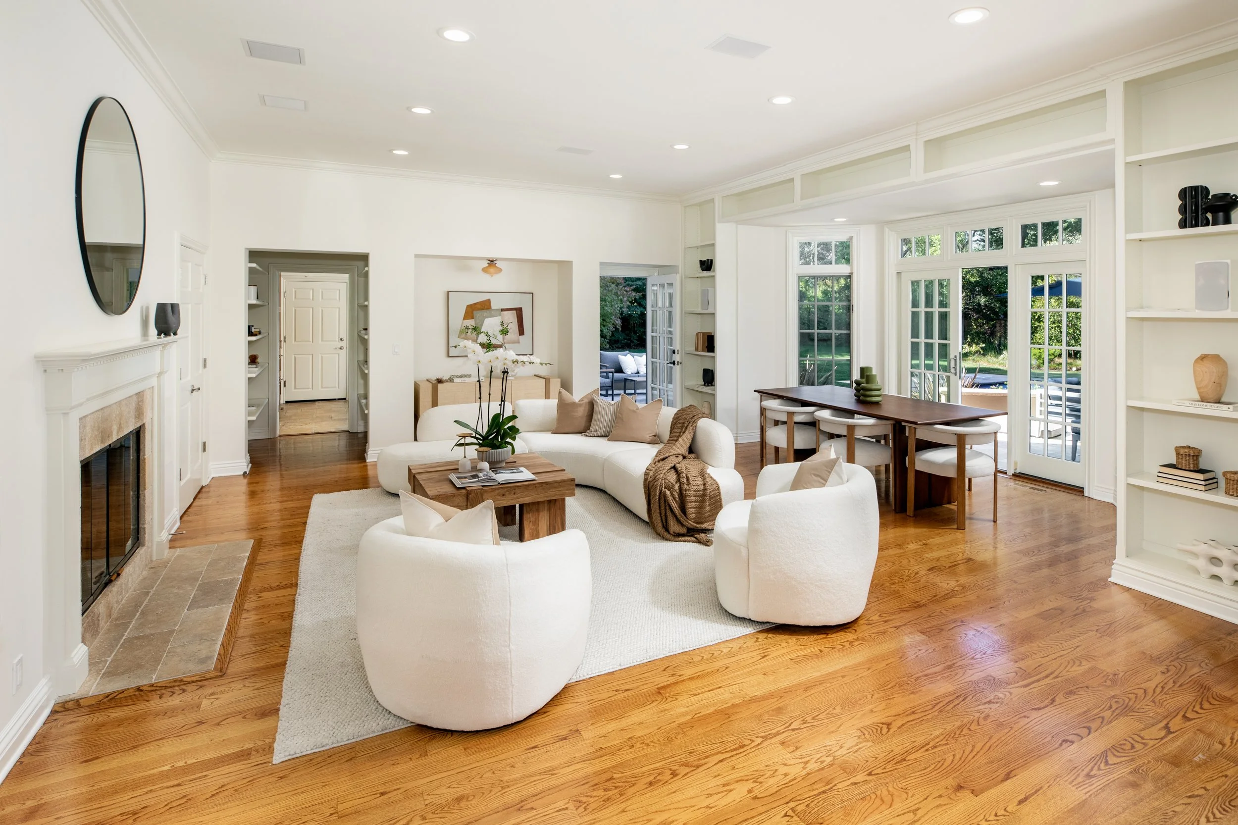 Bright living room with white walls, hardwood floor, plush white sofa, wooden coffee table, and large windows showing an outdoor patio.