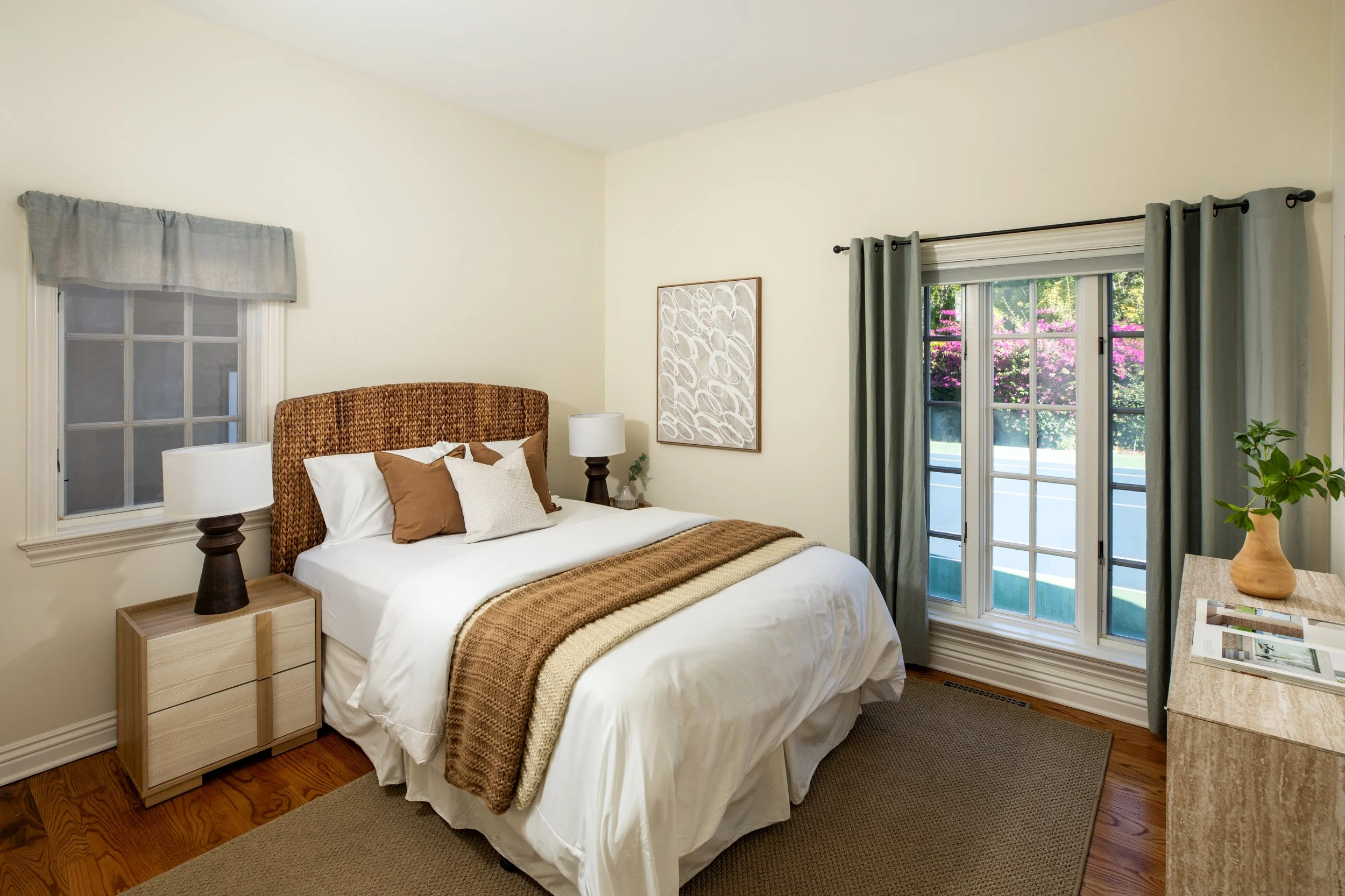 A bedroom with a bed, nightstands, lamps, and windows showing pink flowering bushes outside.