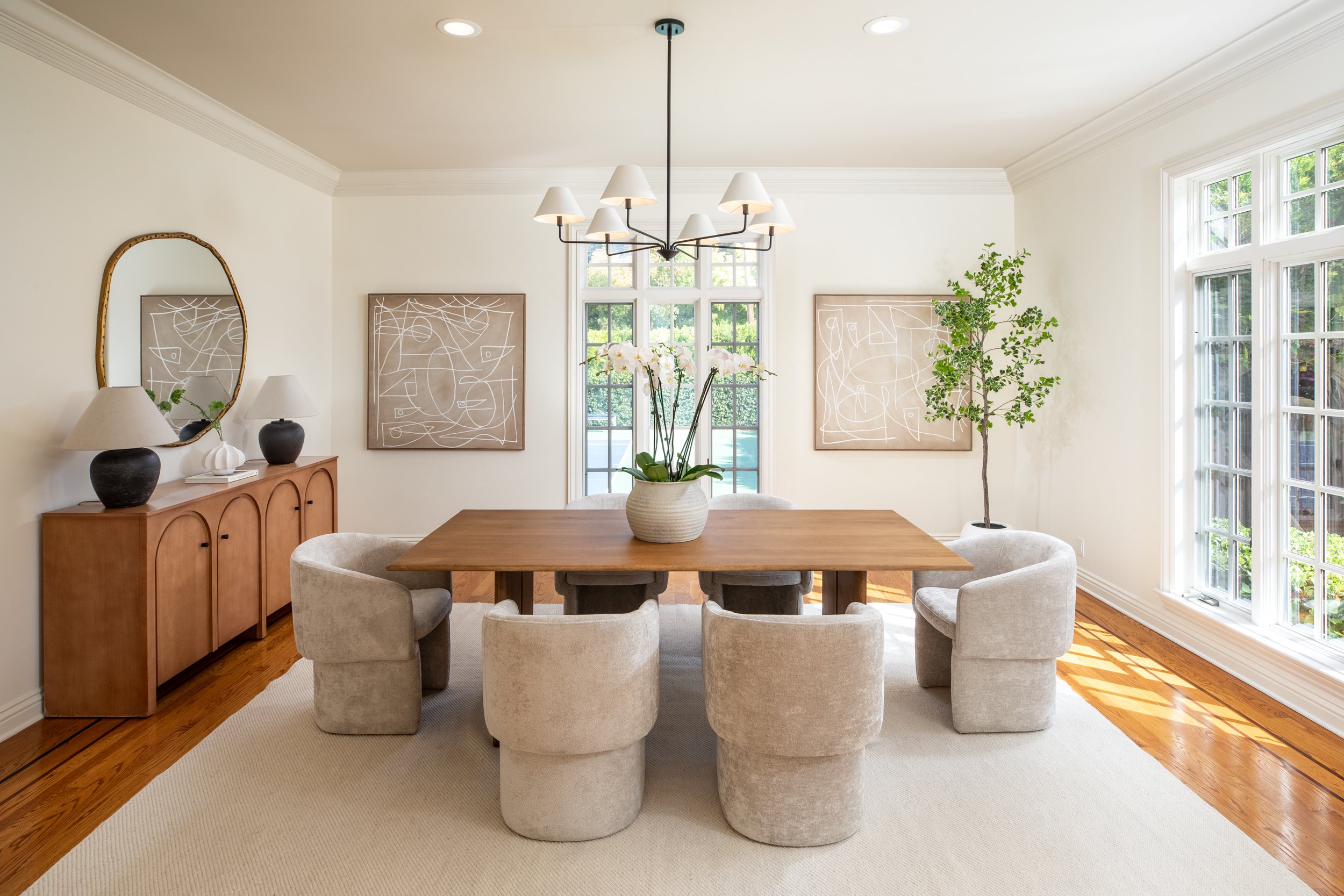 A dining room with a wooden table surrounded by six beige upholstered chairs. A large ceramic vase with white orchids sits on the table. Behind, a sideboard with two black lamps and a mirror. Two abstract art pieces hang on the wall, and large window