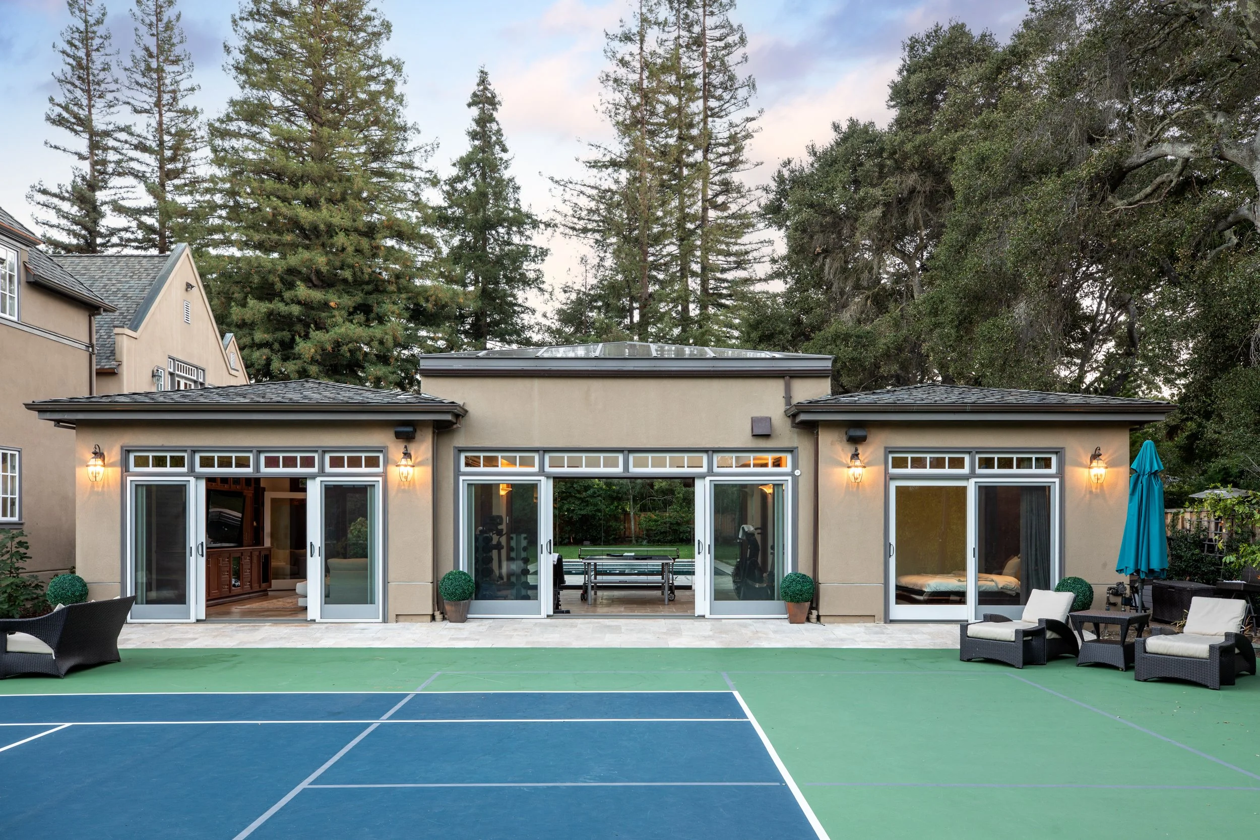 Backyard with tennis court, patio furniture, house with large windows, trees in the background, and outdoor lights.