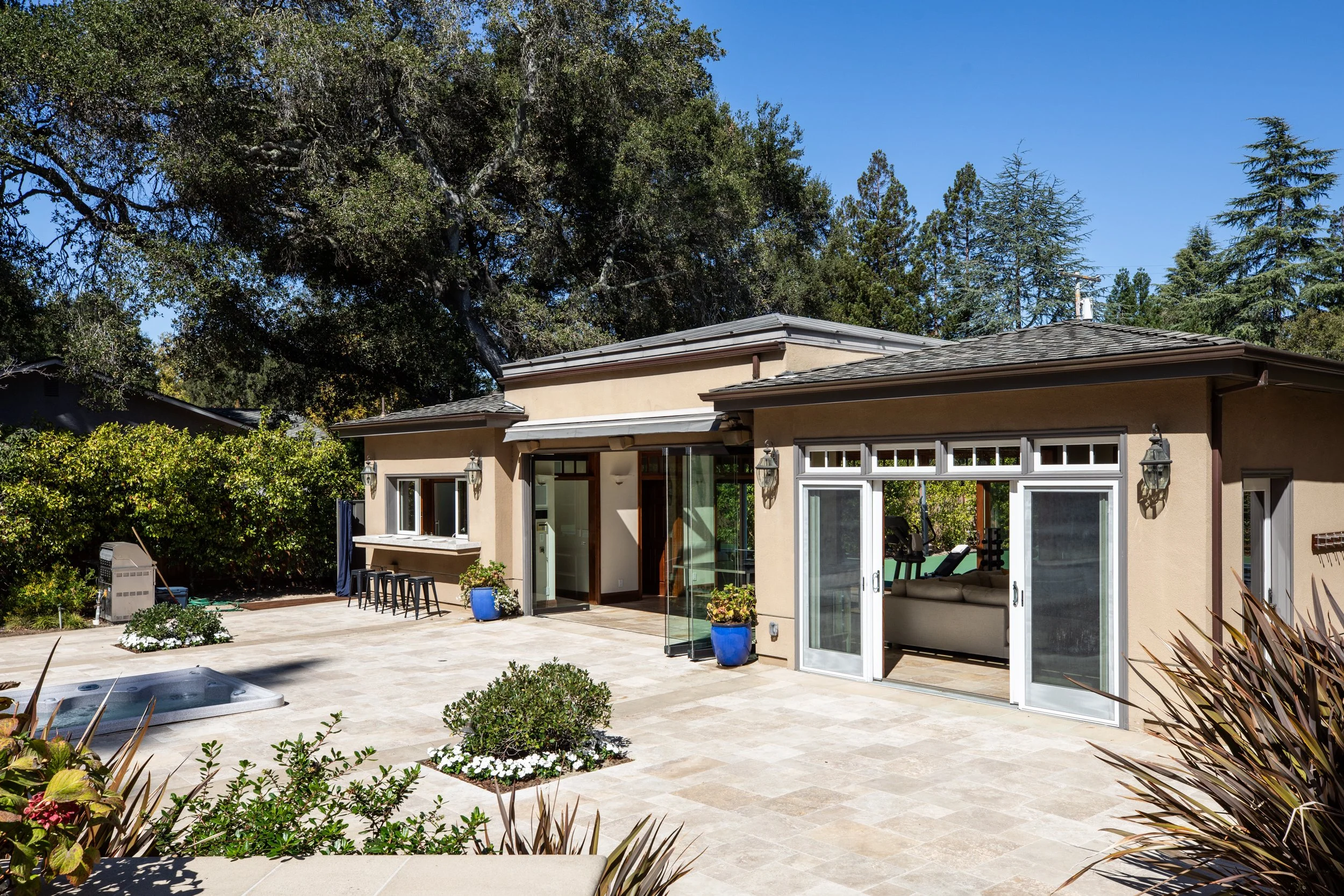 The image shows the backyard patio of a house with a large tree overhead. The house has beige stucco walls and a sliding glass door leading to the patio. The patio has chairs, potted plants, and a hot tub, with greenery and bushes surrounding the are
