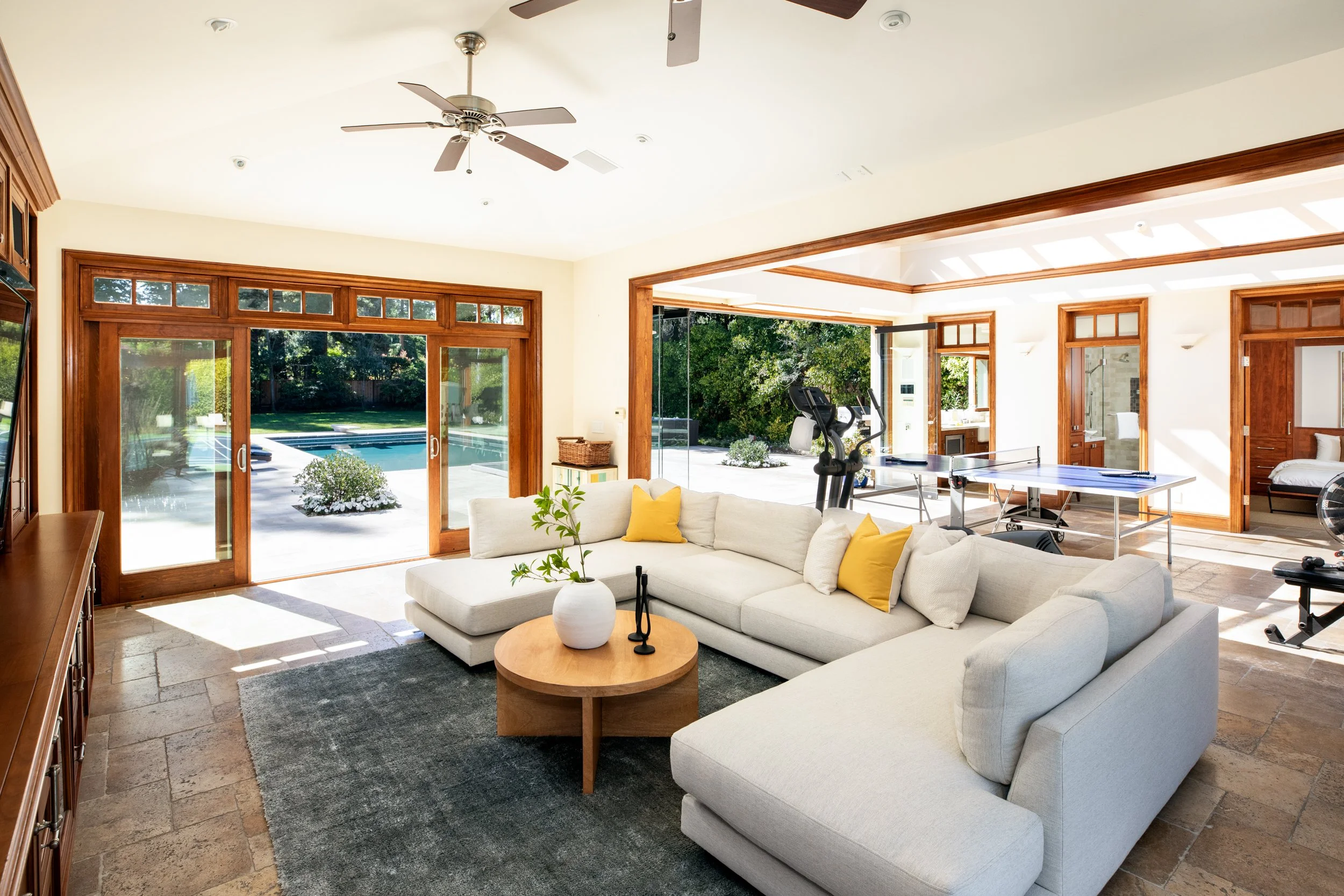 Living room with large glass doors leading to backyard with pool, beige walls, wooden trim, white sectional sofa with yellow pillows, indoor exercise bike, ping pong table, and decorative plants.