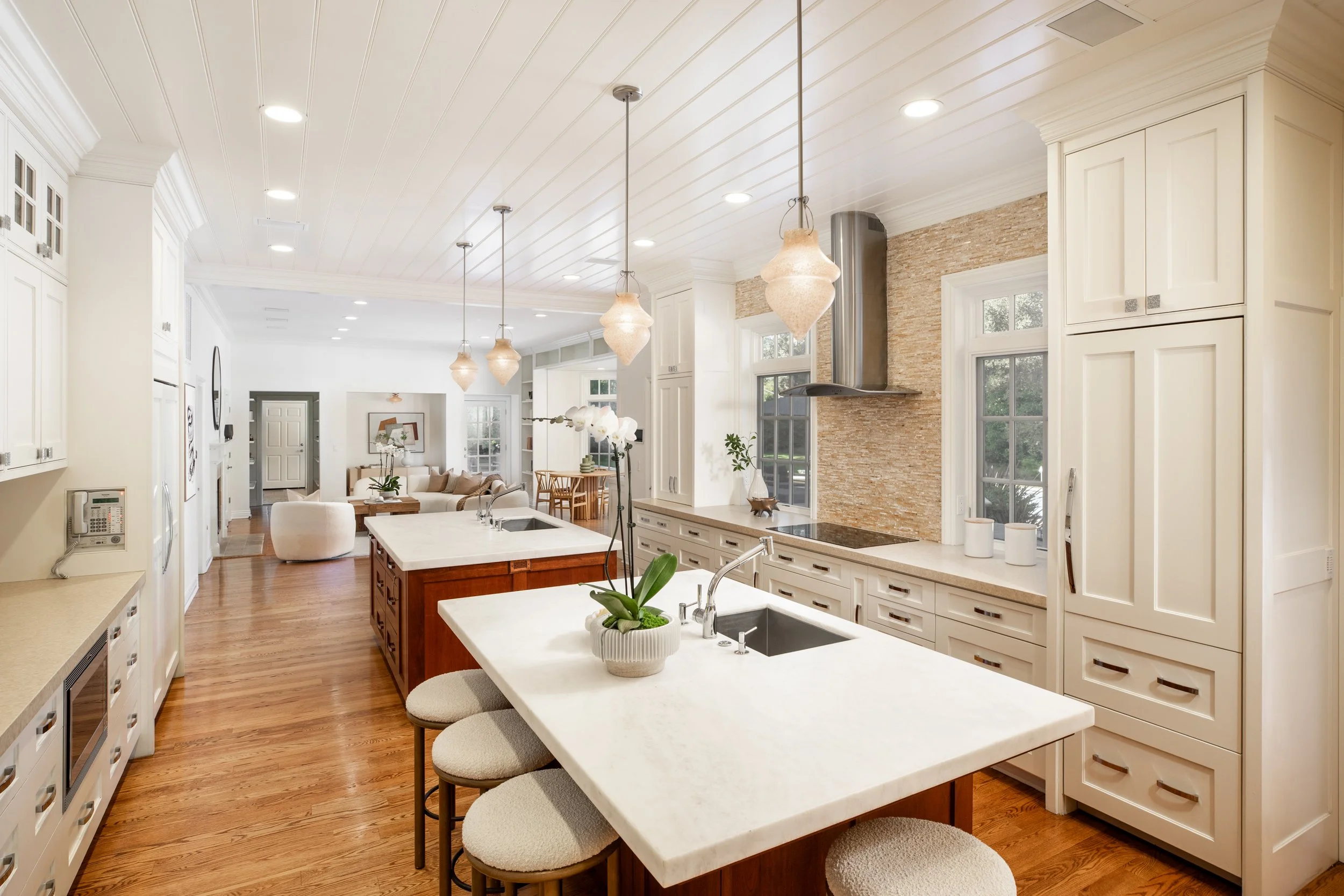 A spacious, modern kitchen with white cabinetry, a large island with a white countertop, and pendant lights hanging from the ceiling. The kitchen overlooks a living area with a sofa and large windows letting in natural light.