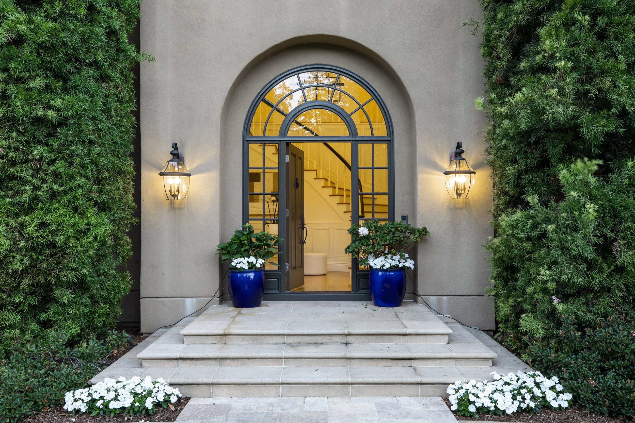Front entrance of a house with gray door and transparent yellow glass, flanked by two blue planters with white flowers, and illuminated by two wall-mounted lanterns, surrounded by green bushes.