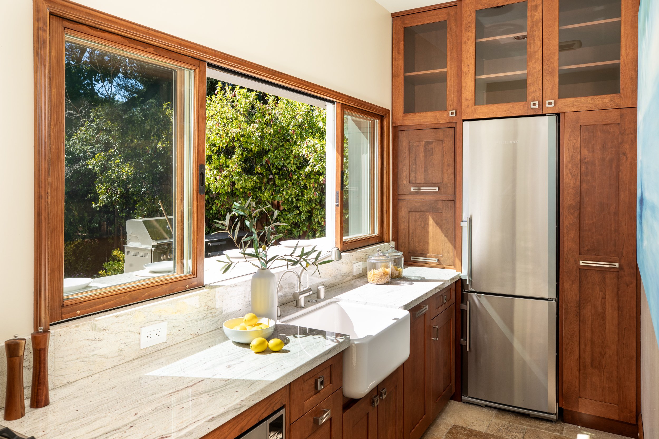 Kitchen with wooden cabinetry, a large window showing greenery outside, a white farmhouse sink, and a stainless steel refrigerator.
