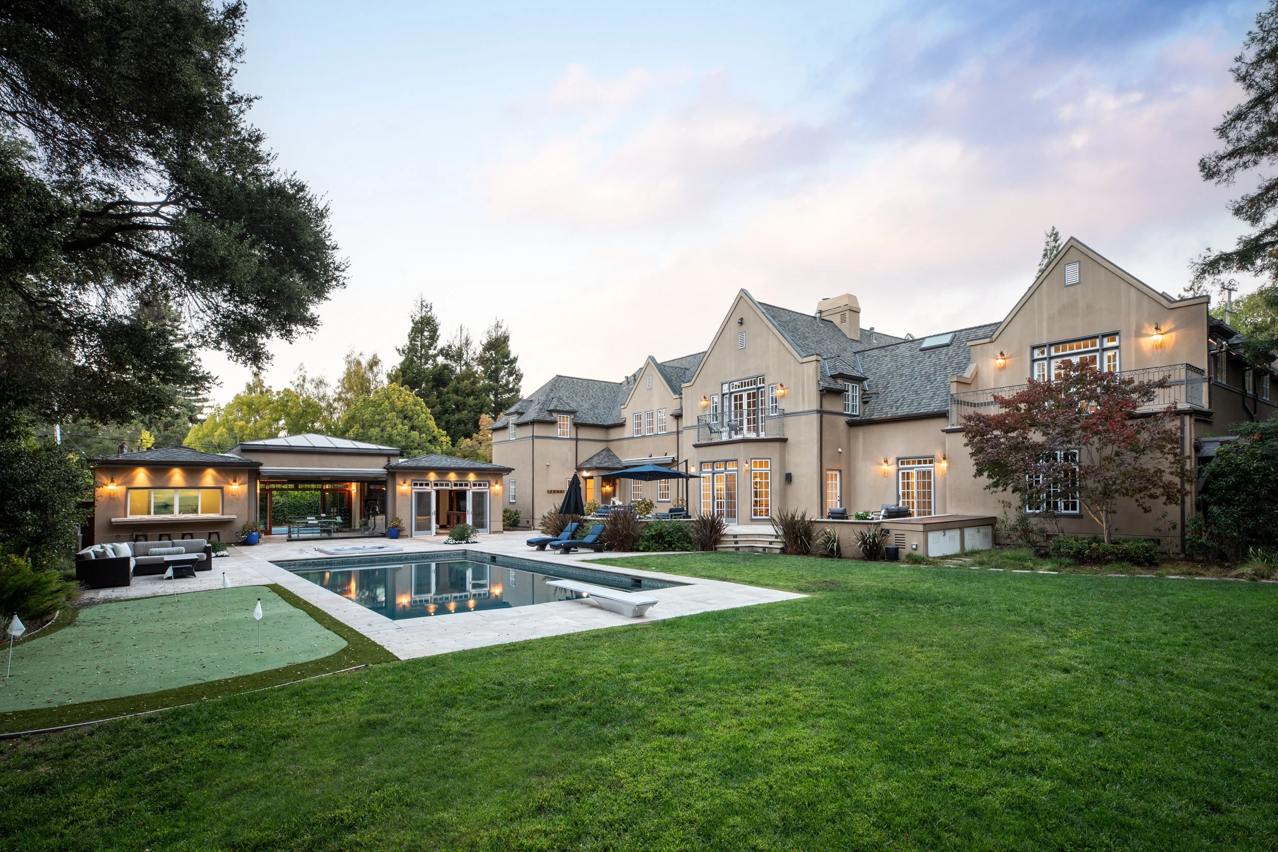 Luxury house with a large backyard featuring a swimming pool, outdoor seating area, and surrounded by trees and greenery, during early evening with lights on