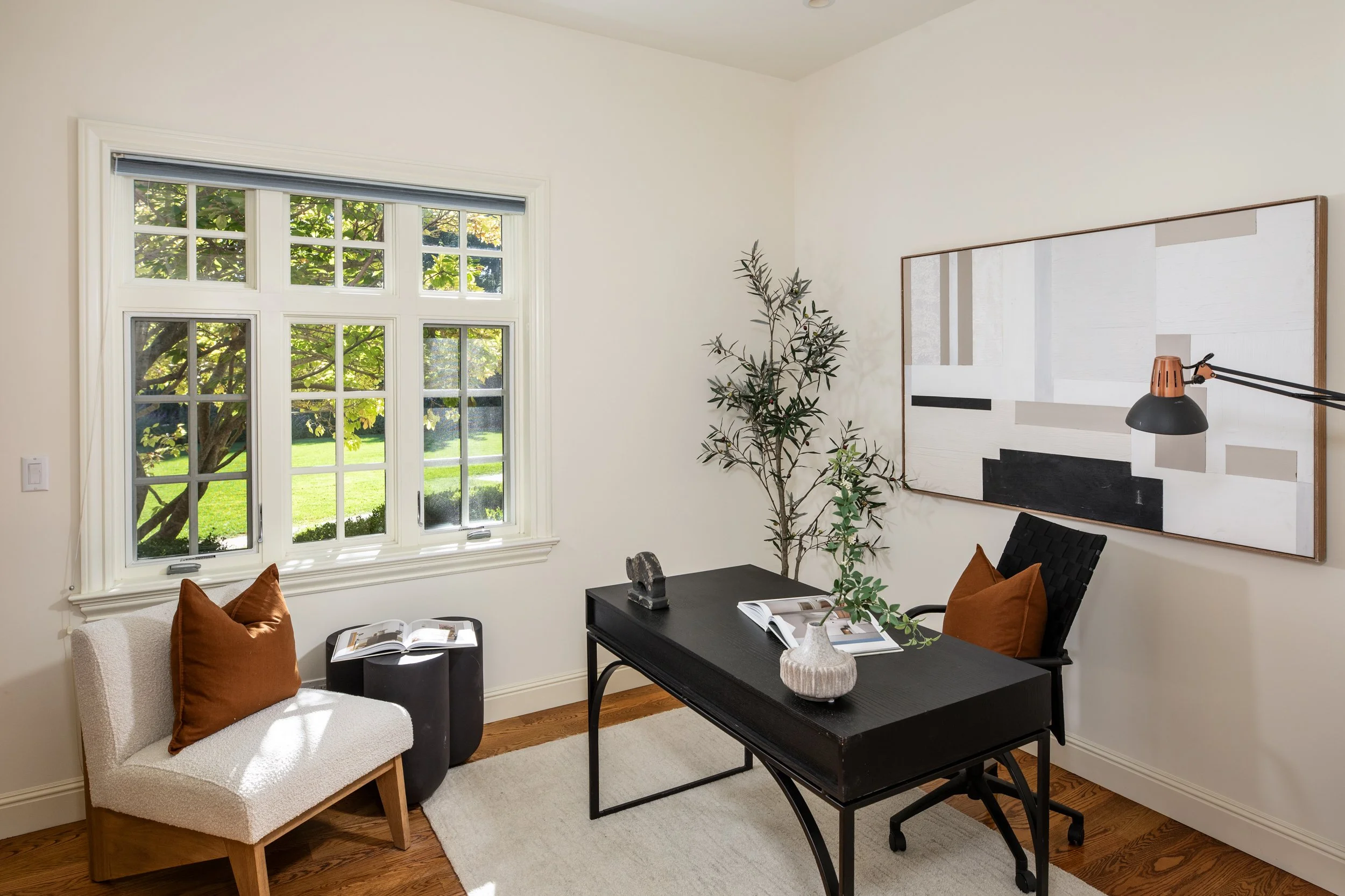 Room with large window, white walls, abstract wall art, black desk with open books, beige armchair with brown cushion, black chair with brown cushion, potted plant.