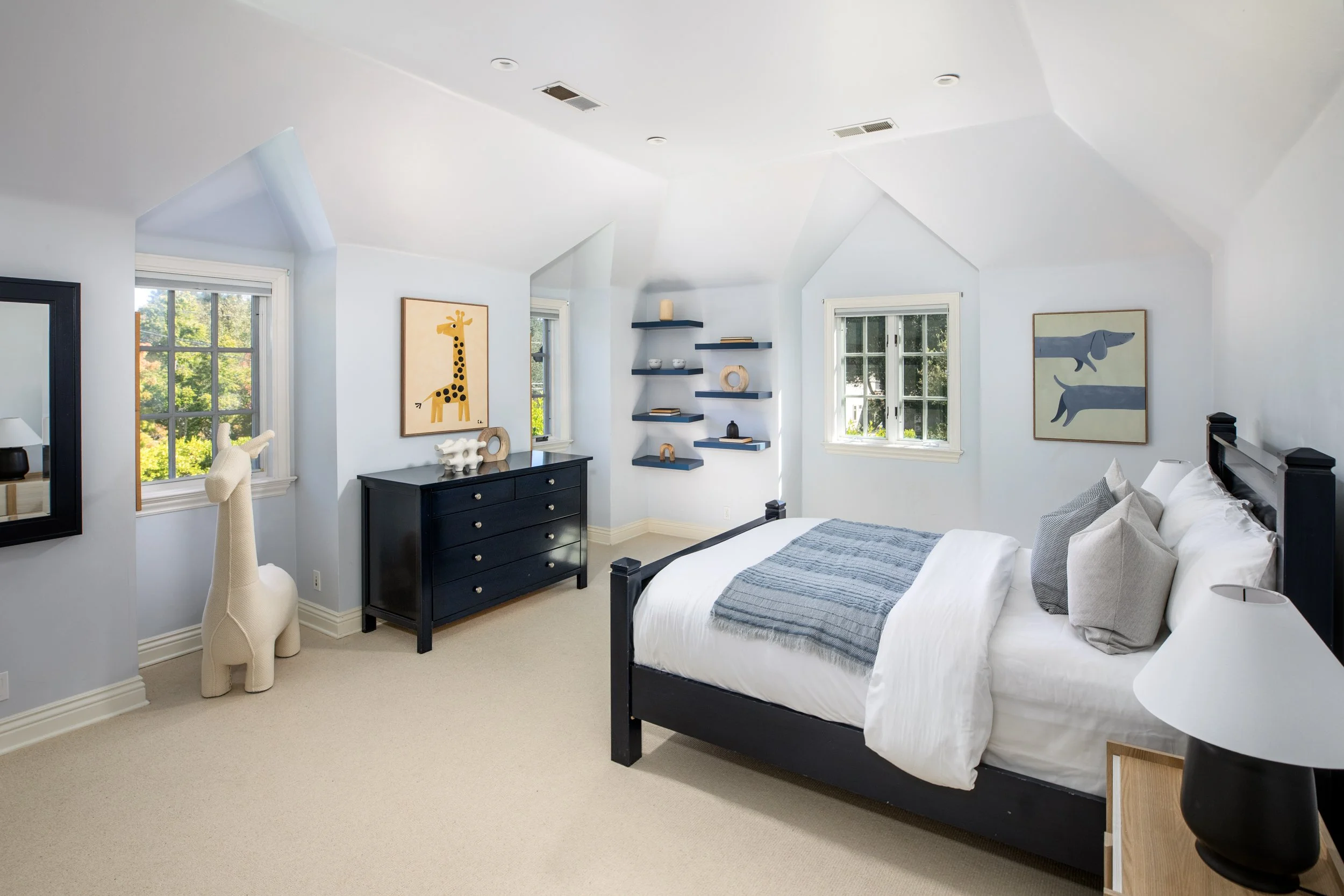 A bedroom with blue walls, a black bed frame with white bedding, a black dresser, and framed animal artwork, with windows showing trees outside.