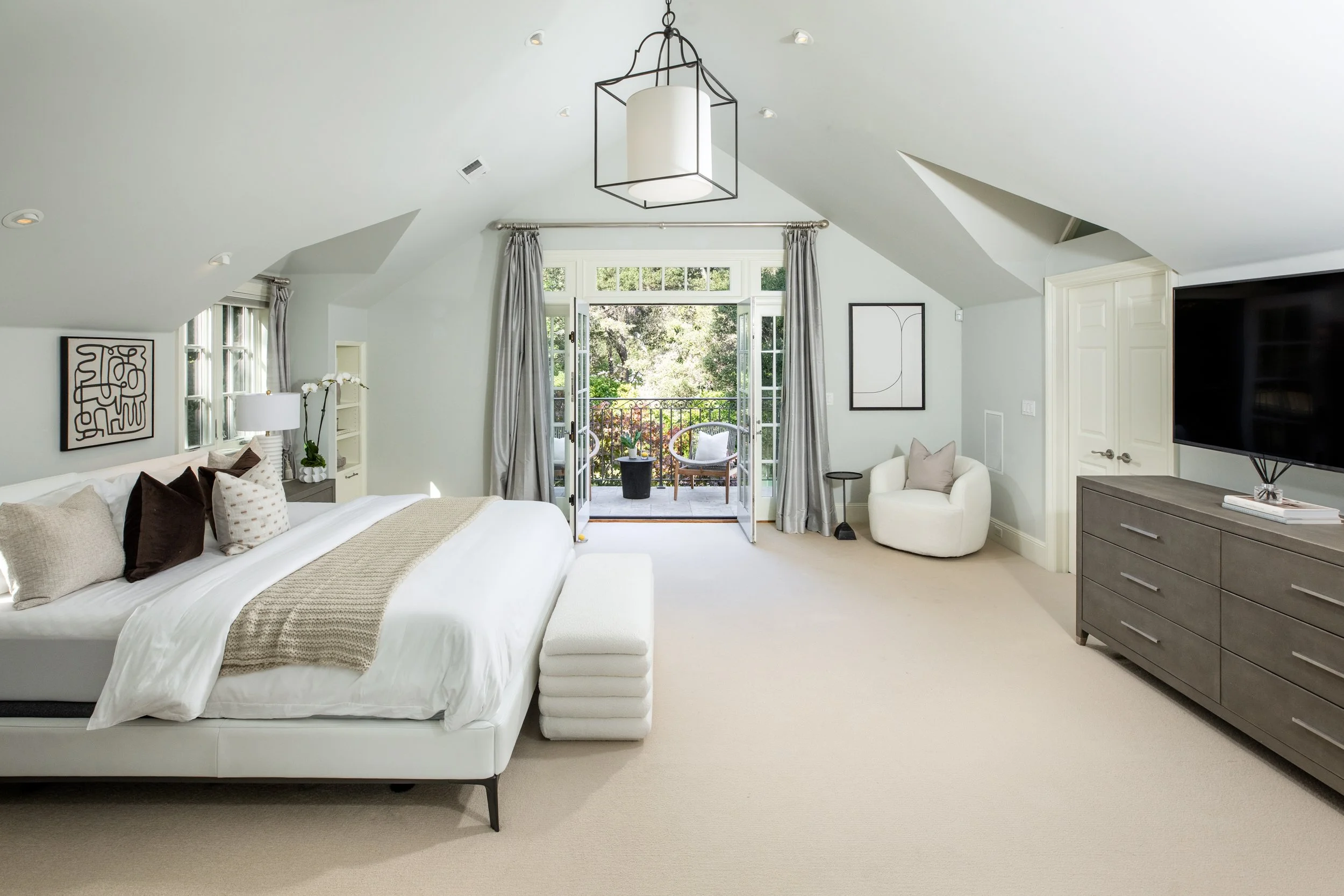 A spacious, well-lit bedroom with a large bed and an open balcony door showing greenery outside. The room contains a white armchair, a gray dresser, wall art, and a ceiling light fixture.