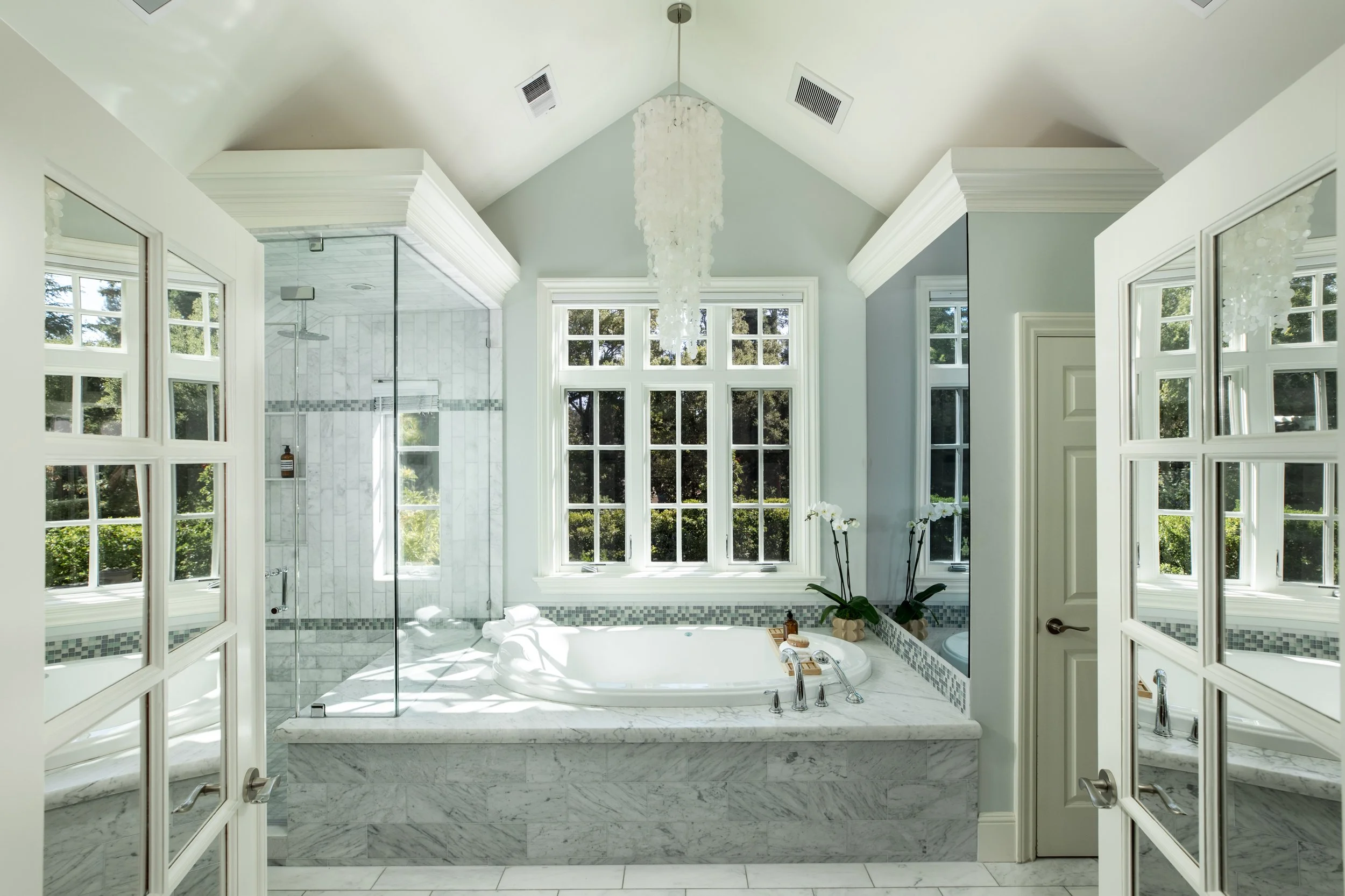 Bright bathroom with large windows, a corner bathtub, and a walk-in shower. White and gray marble finishes, a chandelier, and decorative orchids.