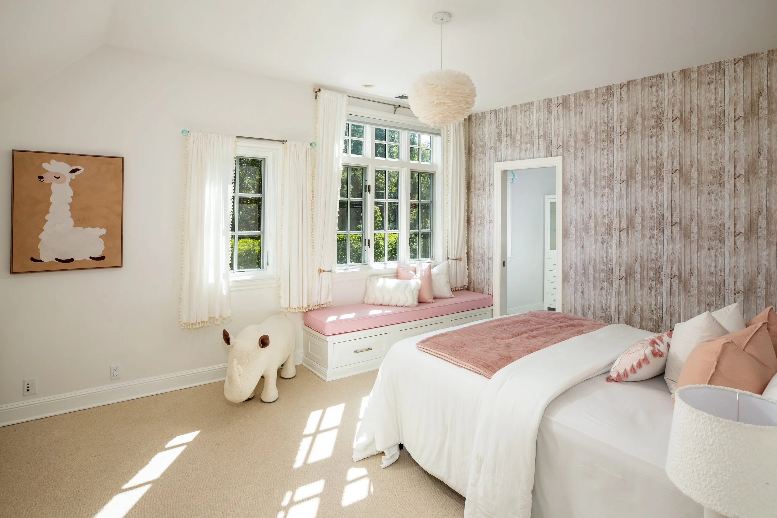Bright bedroom with large windows, white curtains, pink and white bedding, beige carpet, wall art of a llama, and a plush llama decor near the window.