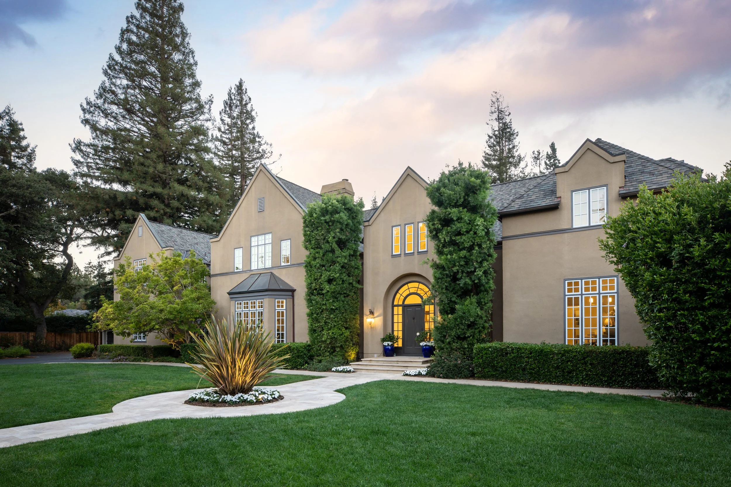 Front view of a large beige house with multiple gable roofs, surrounded by green trees, a well-maintained lawn, and a curved stone pathway leading to the front door, which is illuminated and flanked by potted plants.