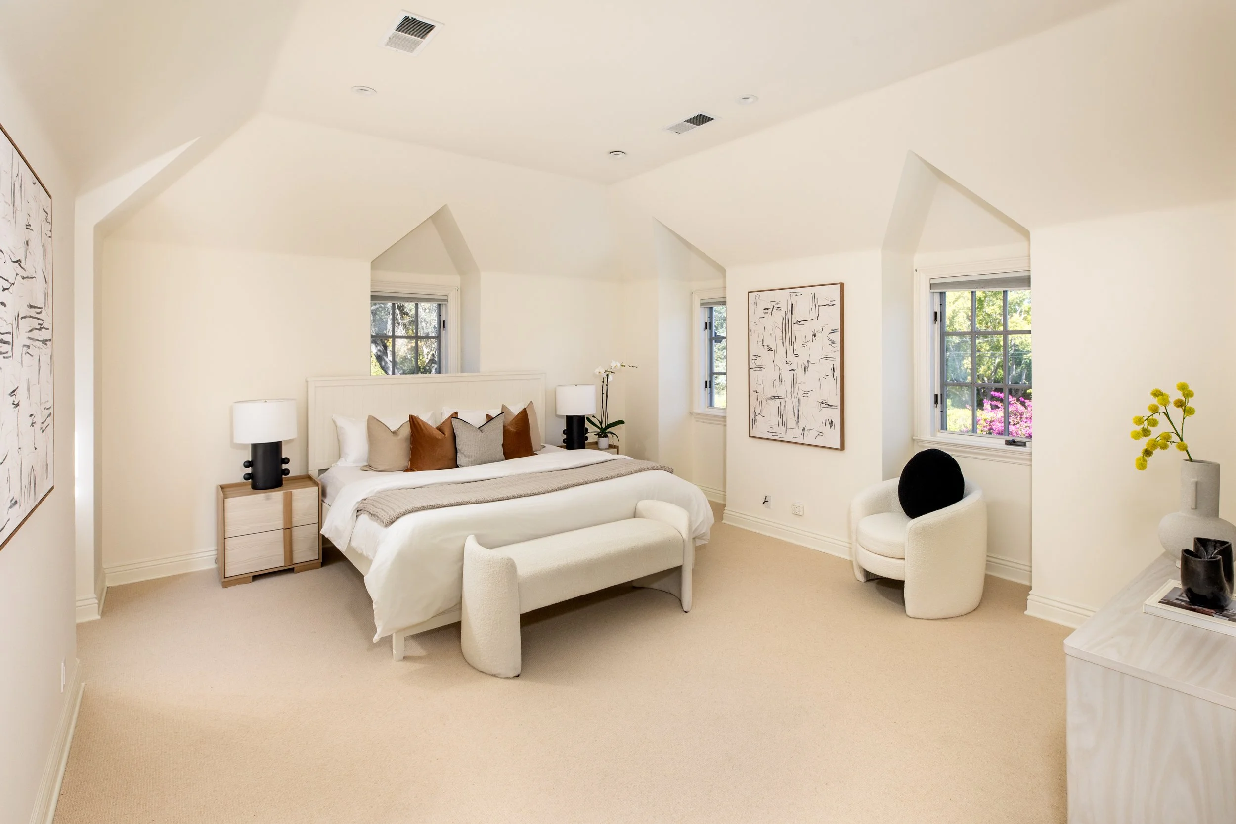 A cozy bedroom with a white bed, beige and brown pillows, two nightstands with white lamps, and artwork on the walls. There are three windows showing greenery outside, and a small white chair with a black cushion near a vase with yellow flowers.