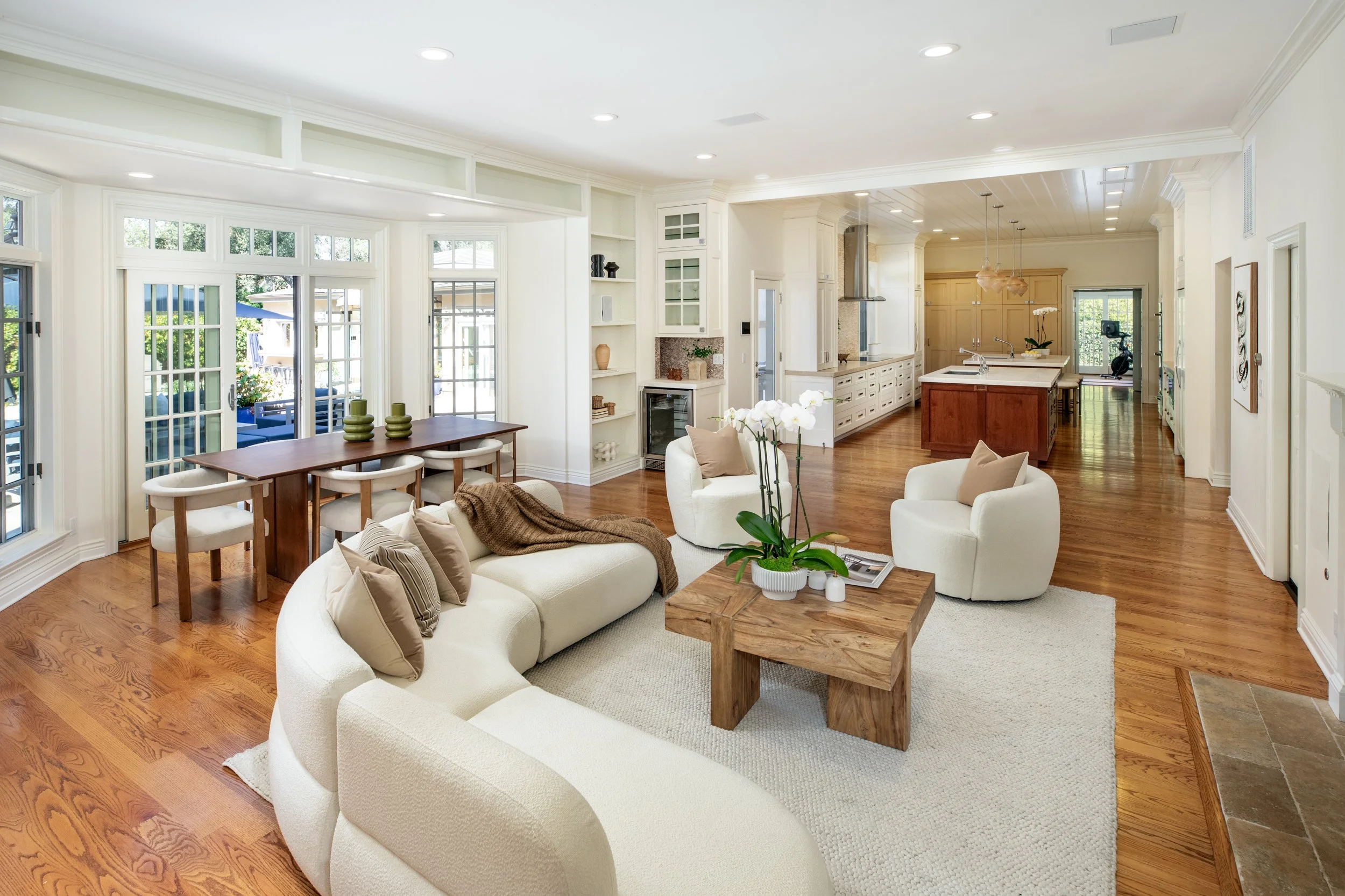 Open concept living room and kitchen with white walls and hardwood floors, featuring cream-colored sofa, round chairs, wooden coffee table, and a kitchen with an island and pendant lights.