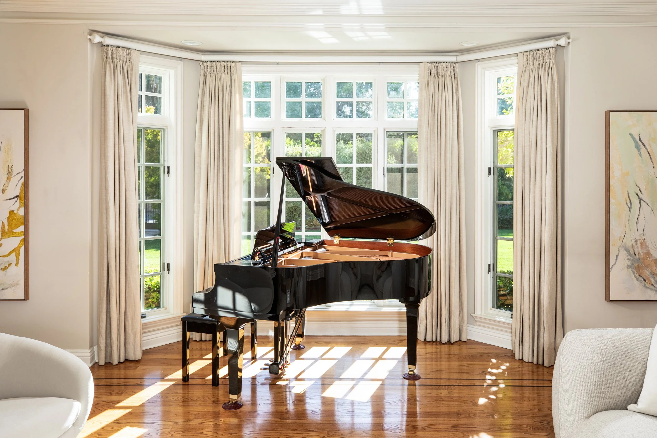 A black grand piano in front of a large window with white trim and beige curtains, sunlight streaming in through the window, and artwork on the walls.