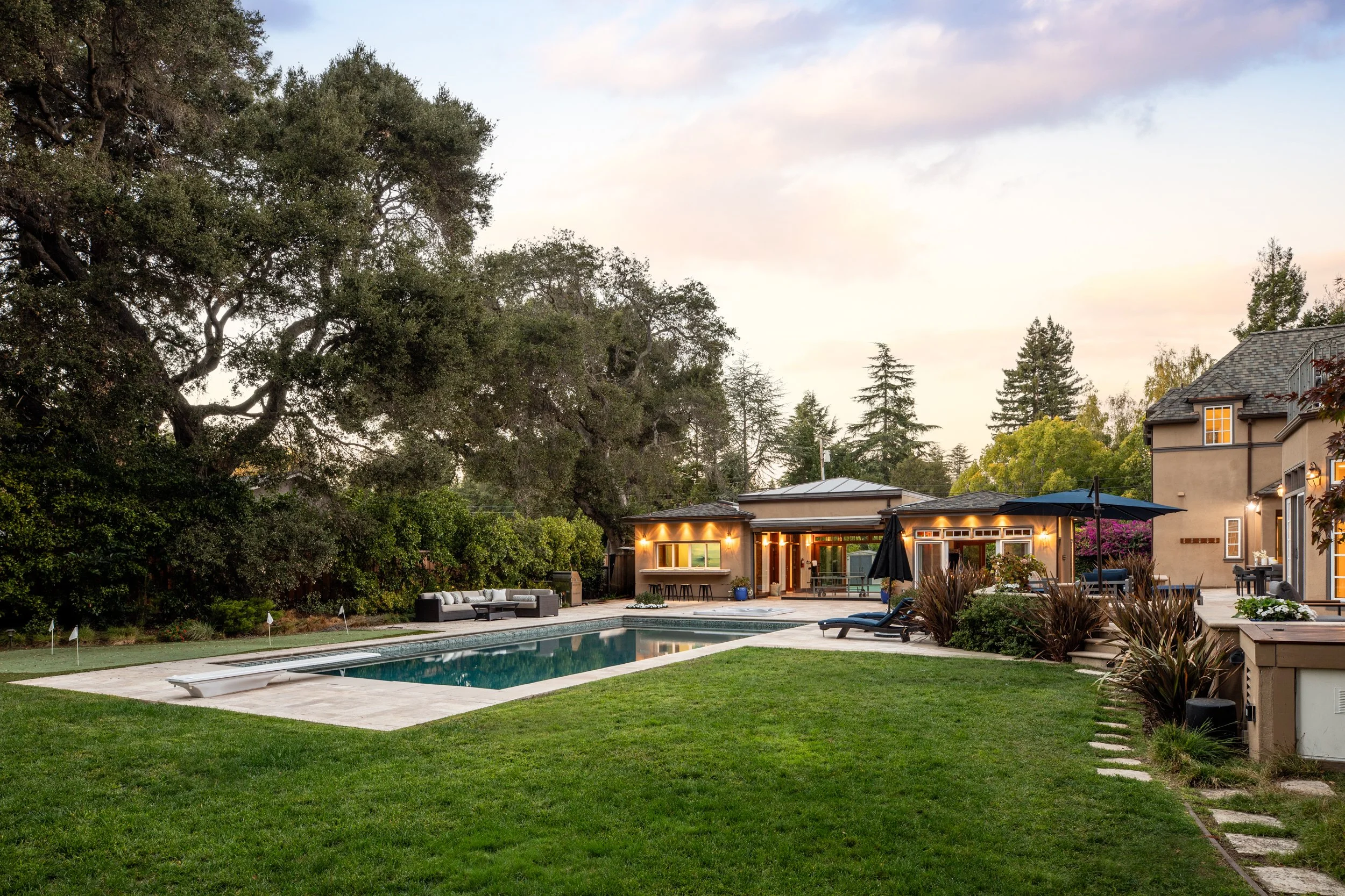 A backyard with a swimming pool, patio furniture, and lush greenery with trees and a multi-story house in the background, during dusk with lights turned on inside