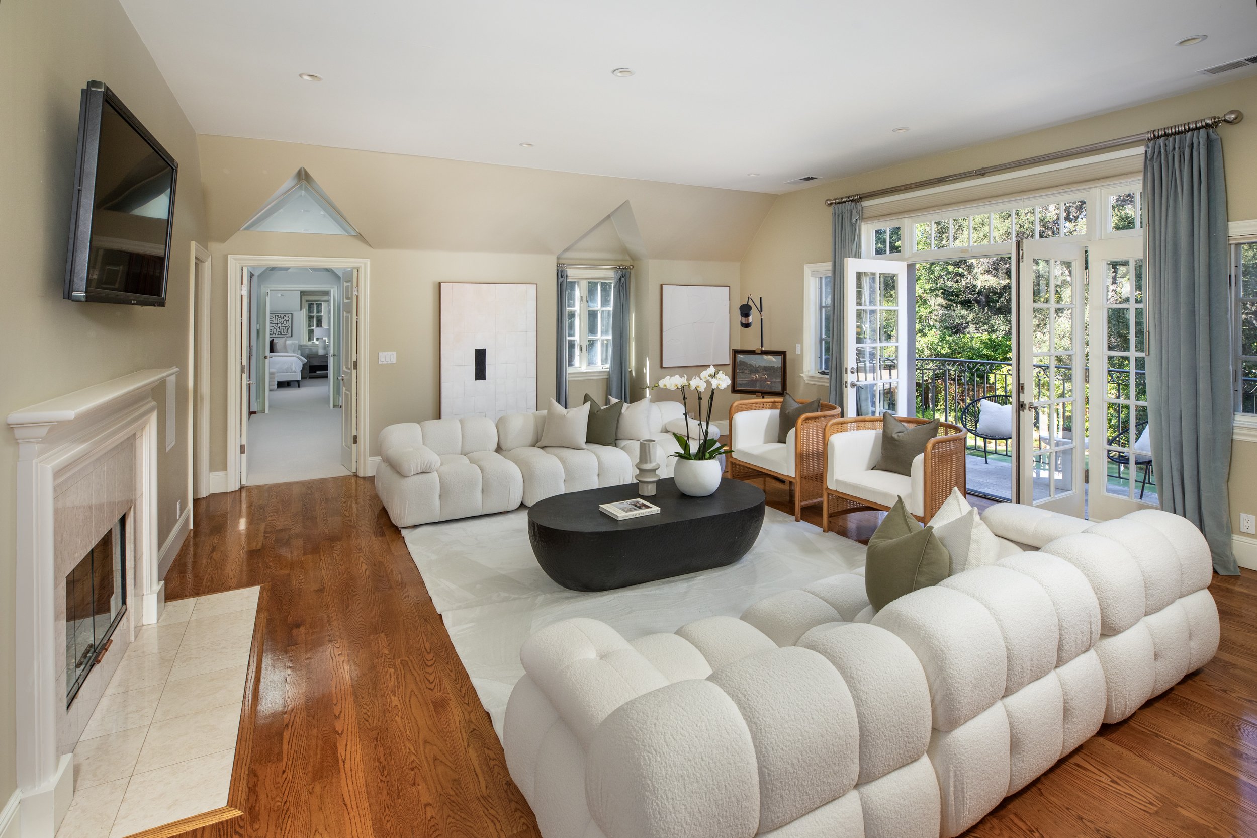 Bright living room with large windows, white upholstered sectional sofa, chairs, black oval coffee table, and wall-mounted TV.