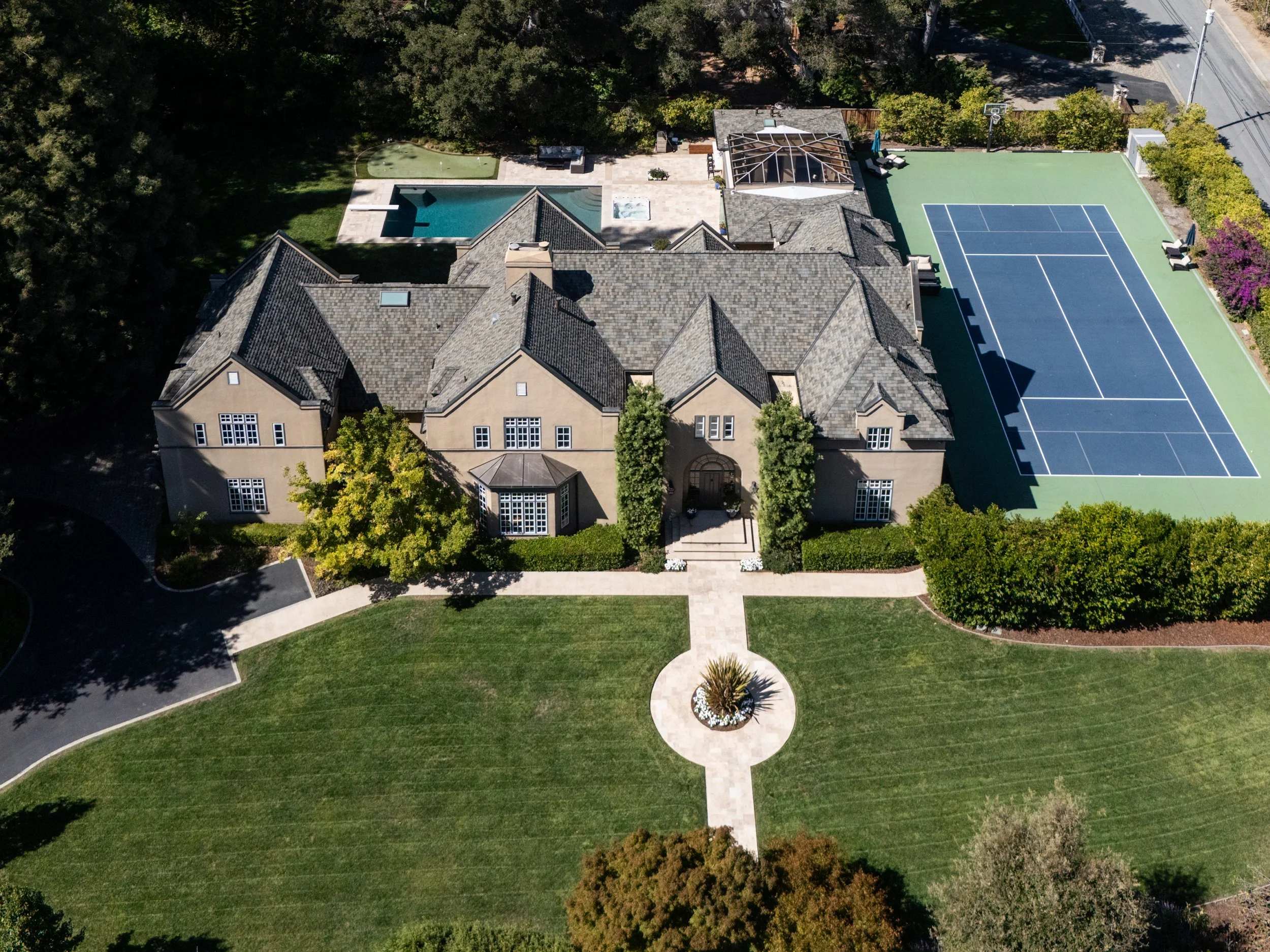 Aerial view of large mansion with swimming pool, tennis court, sidewalk, trees, and road