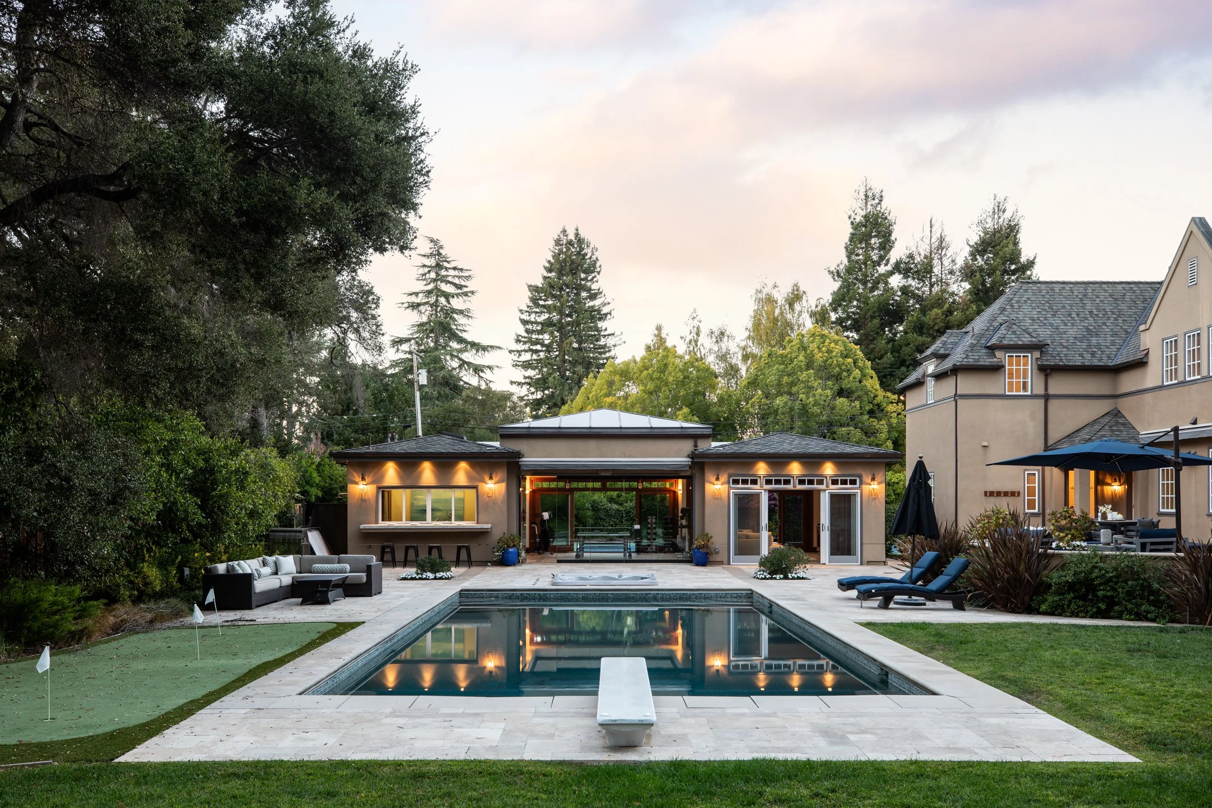 A backyard view of a modern house with large windows and a rooftop deck, surrounded by tall trees, with a swimming pool, outdoor seating area, and a hot tub, during dusk with outdoor lights on.