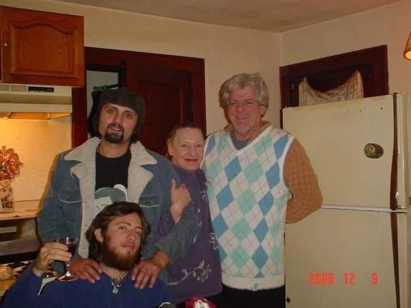 A group of four people posing in a kitchen, three men and one woman, smiling at the camera. One man is seated with a drink, while the others stand behind him.
