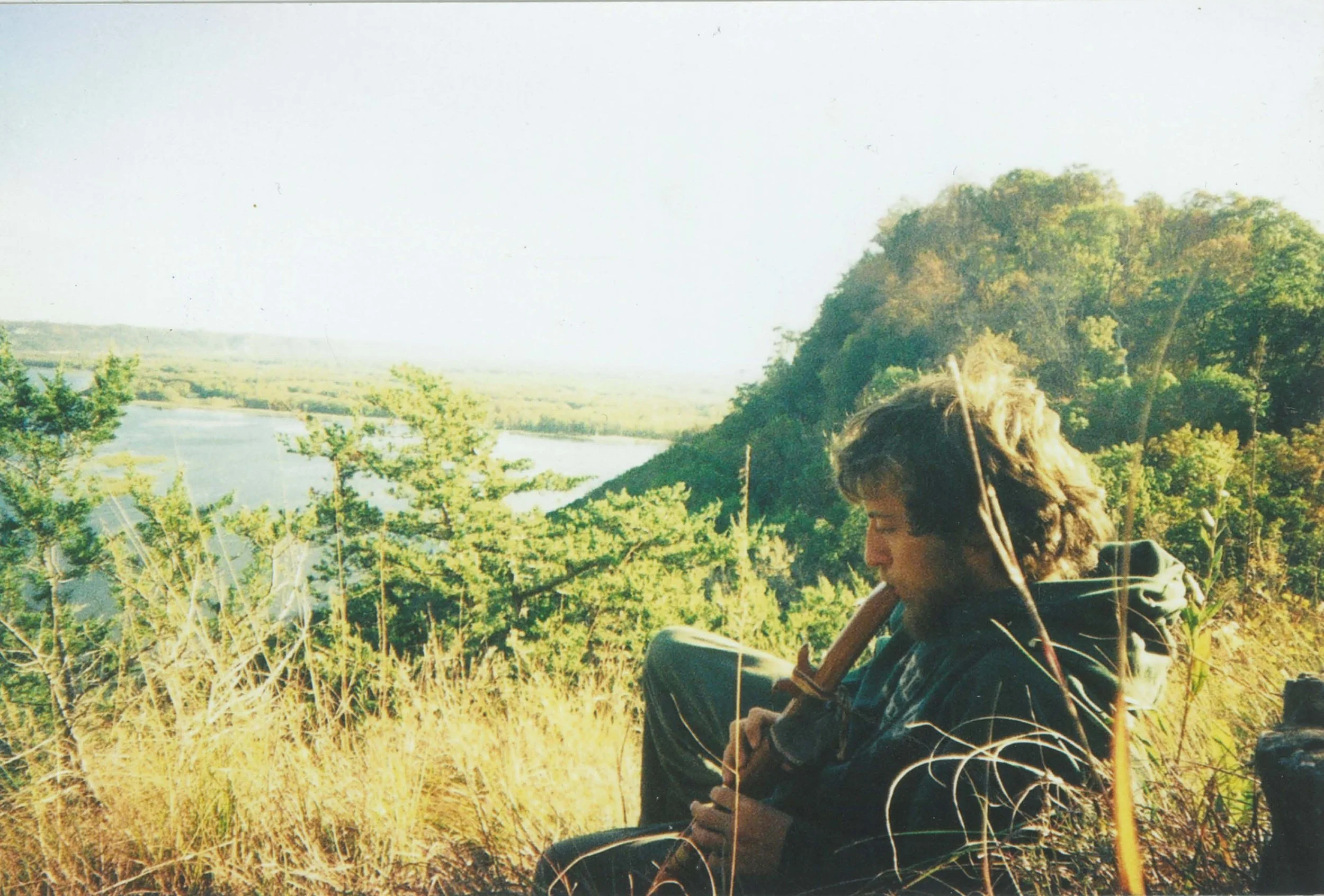 Person sitting outdoors among tall grass and trees, with a river and forested landscape in the background on a bright, sunny day.