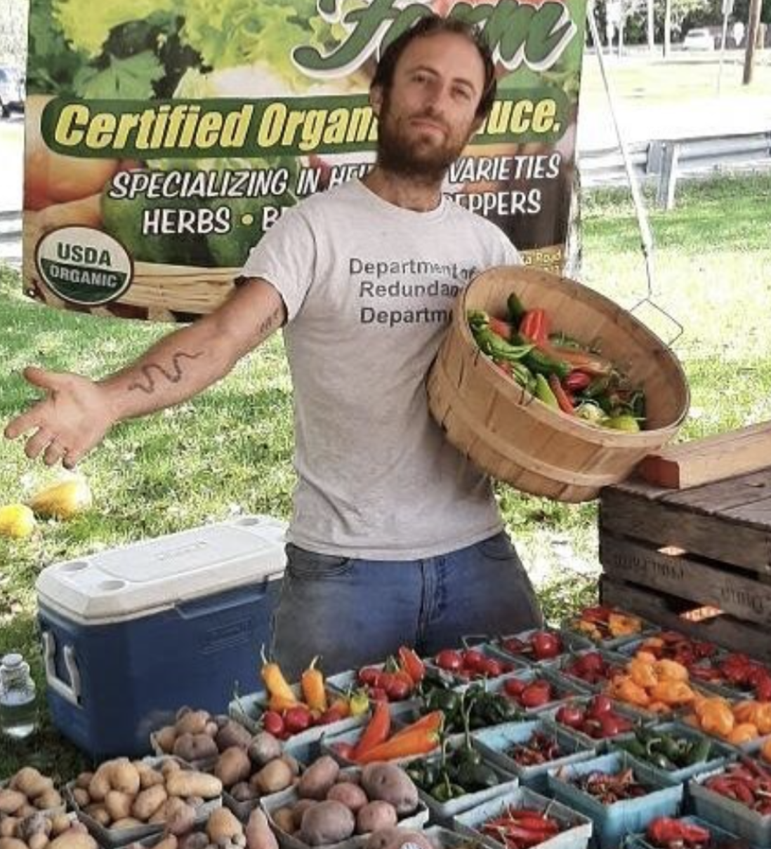 A man at an outdoor farmers market stands behind a table of fresh vegetables, holding a wooden basket filled with peppers and tomatoes. He gestures outward with a welcoming expression, with a banner advertising certified organic produce in the background.