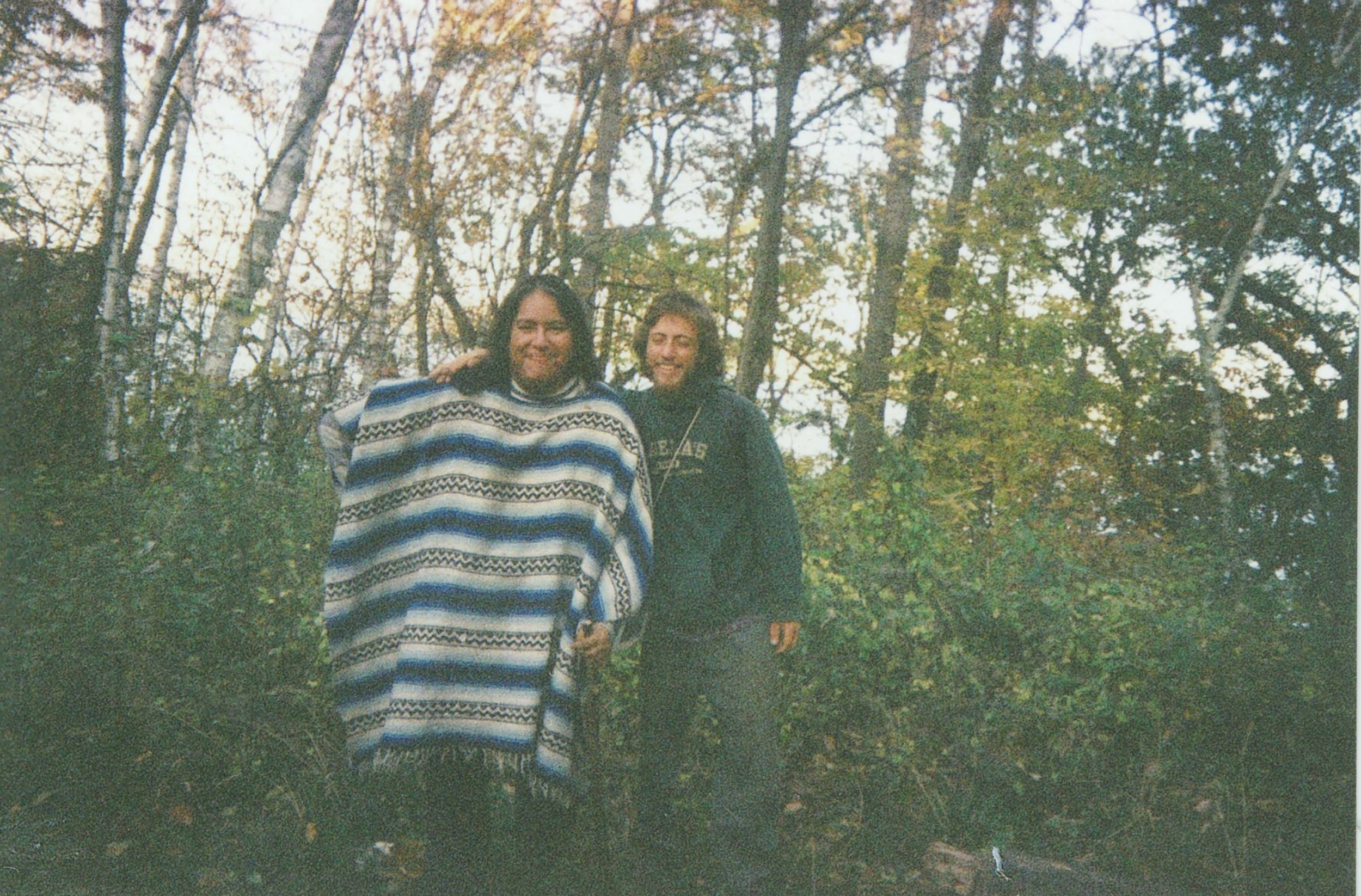 Two people standing outdoors in a wooded area during fall, smiling at the camera. One person is wearing a striped blanket or poncho and the other is wearing a dark hoodie.