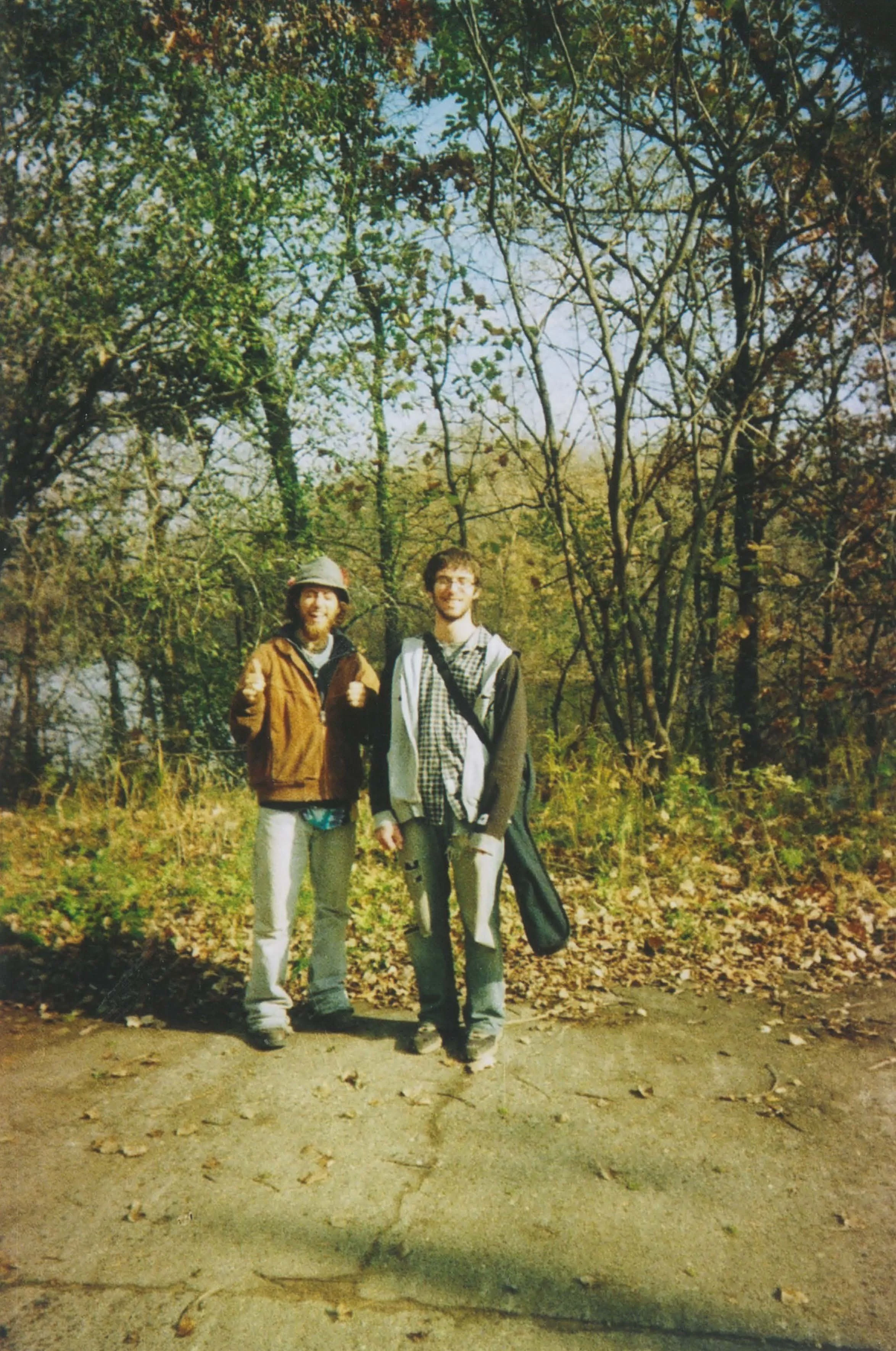 Two men smiling and standing outdoors in a wooded area during fall, dressed casually, with trees and fallen leaves.
