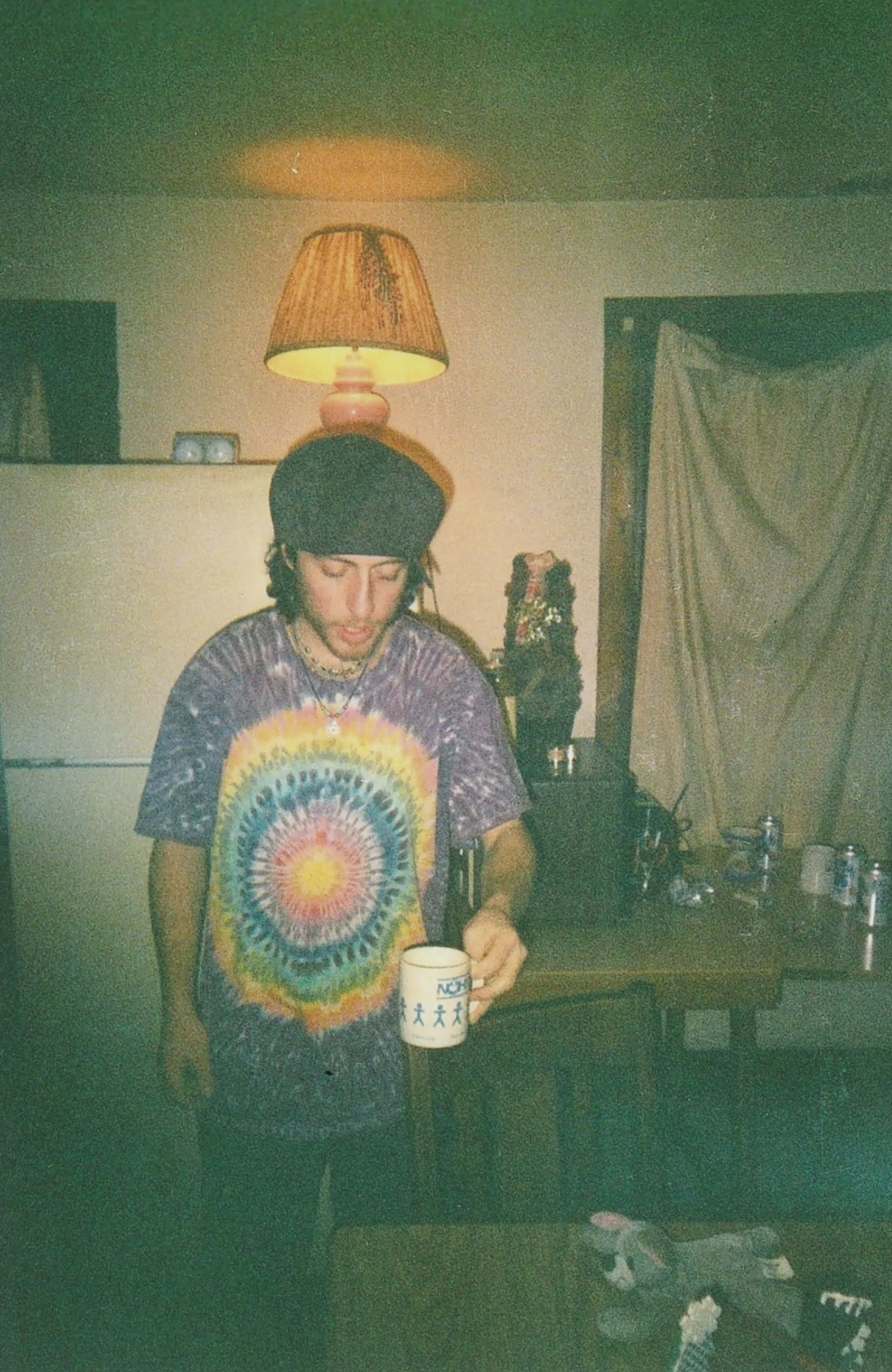 A young man with dark hair and a beanie hat wearing a colorful tie-dye t-shirt, standing in a room with a lit table lamp above his head. He is holding a white mug with a blue design, and there are various bottles and objects on a black table behind h