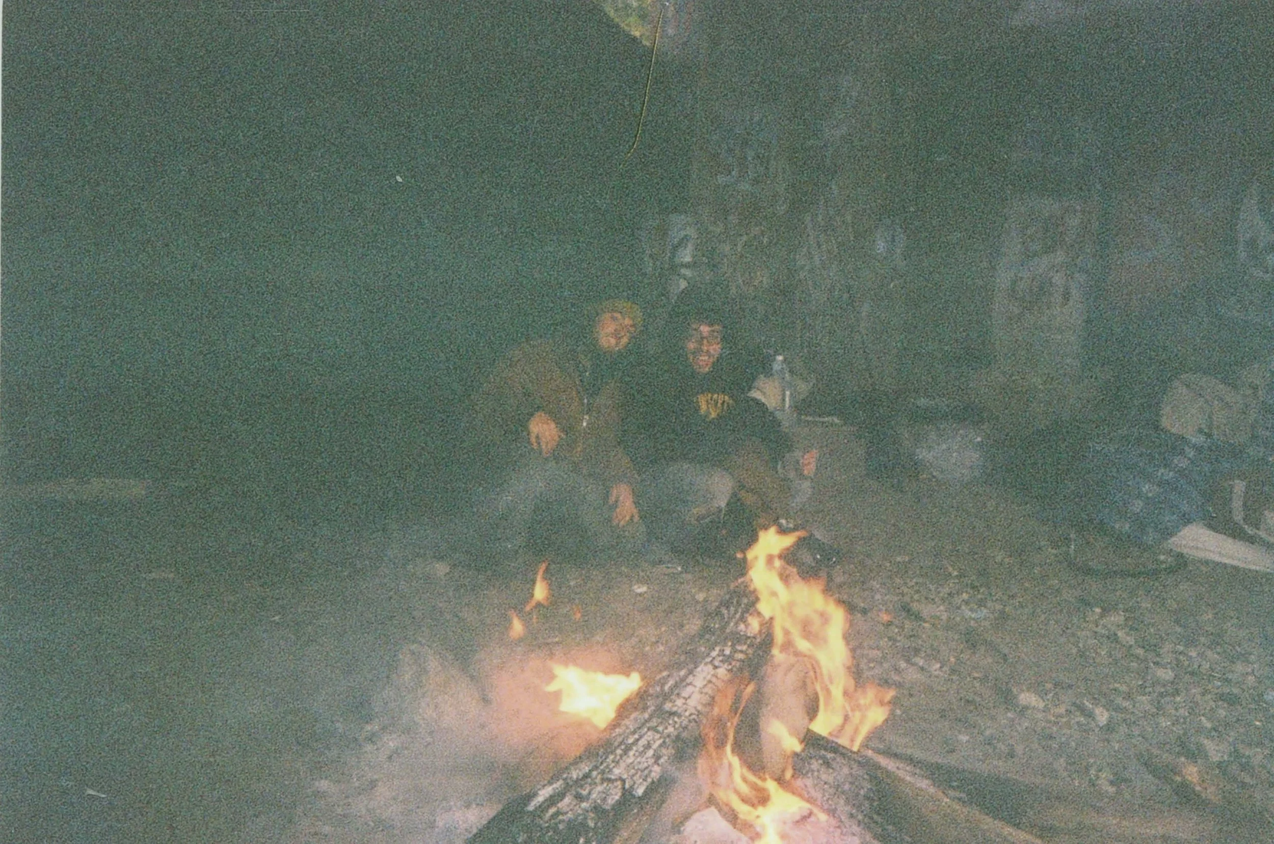 Two people sitting near a campfire at night with trees visible in the background.