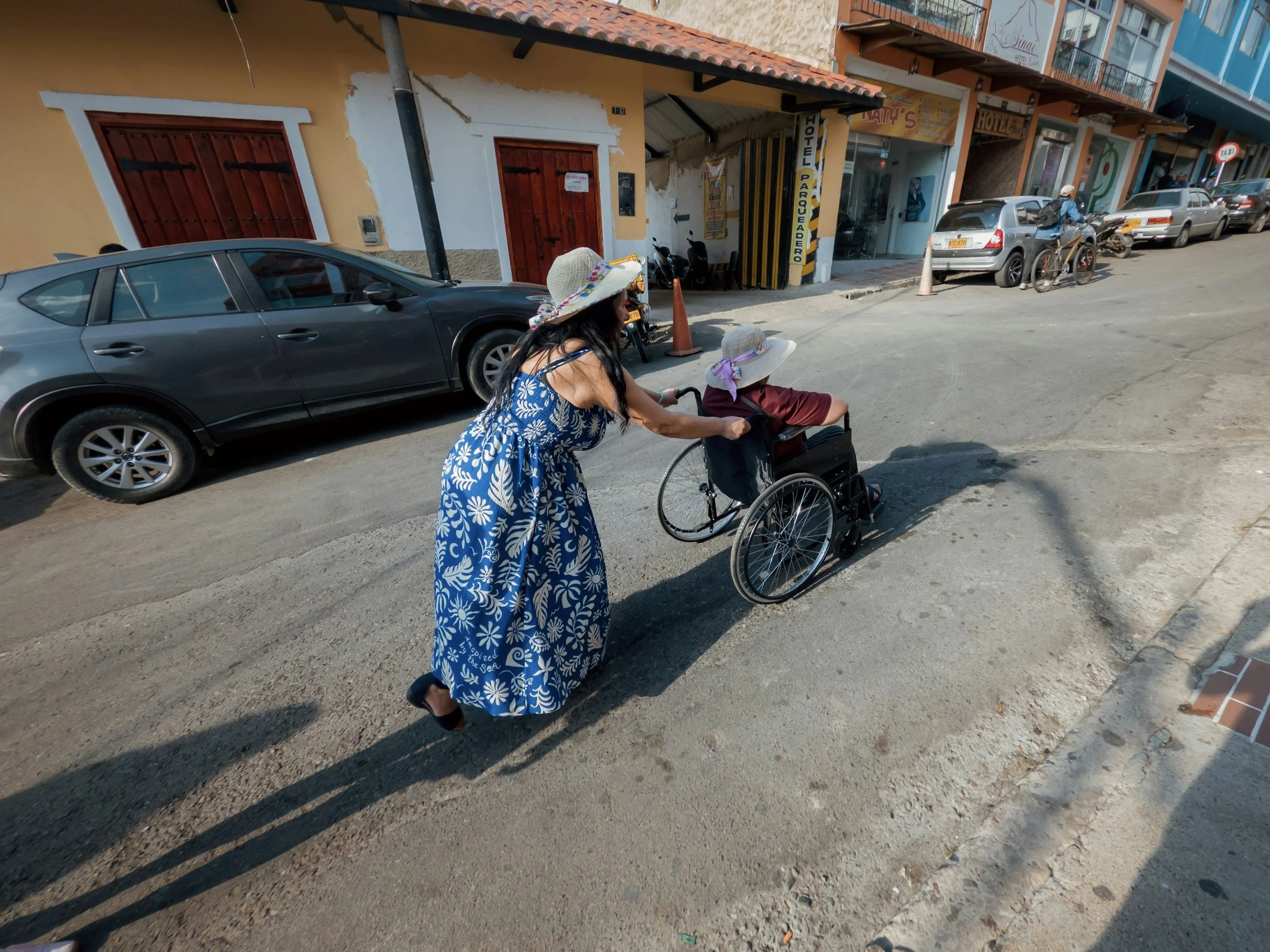 A woman in a floral dress and a wide-brimmed hat is pushing an elderly person in a wheelchair along a city street. The street is lined with parked cars and buildings, some with signs and balconies.