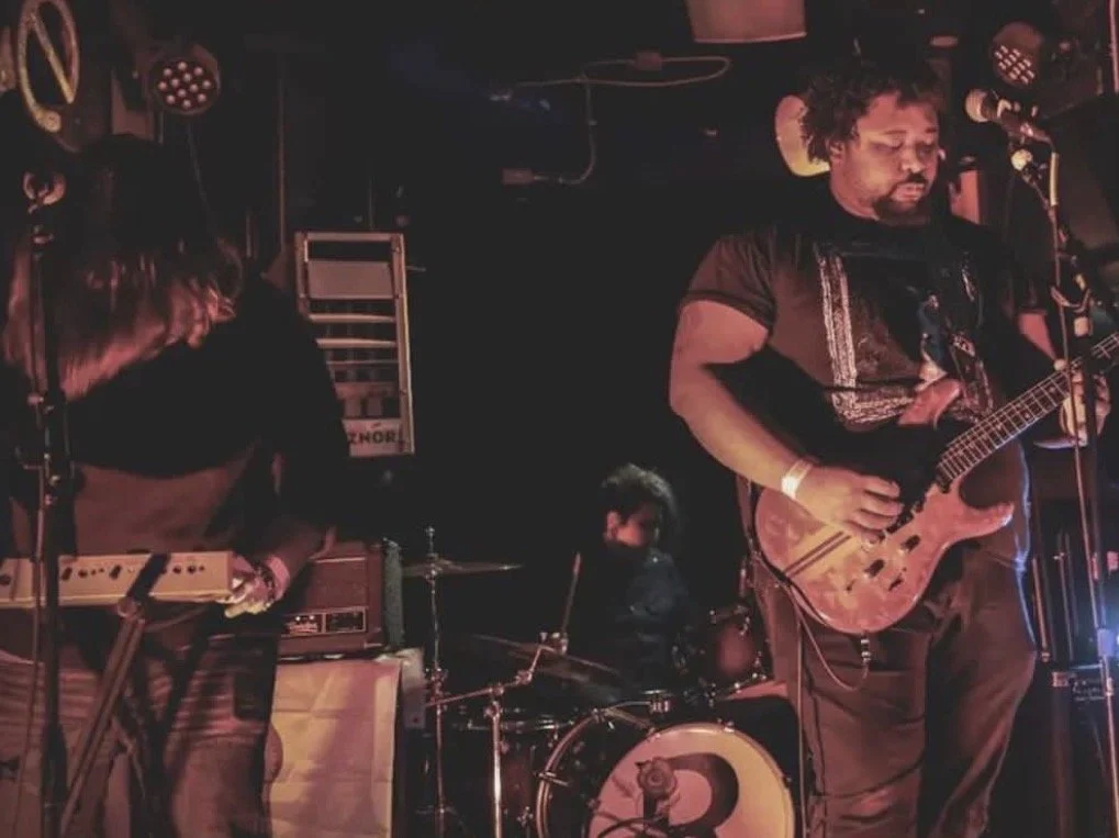A three-member band performing on stage, with a guitarist, a keyboardist, and a drummer in the background.