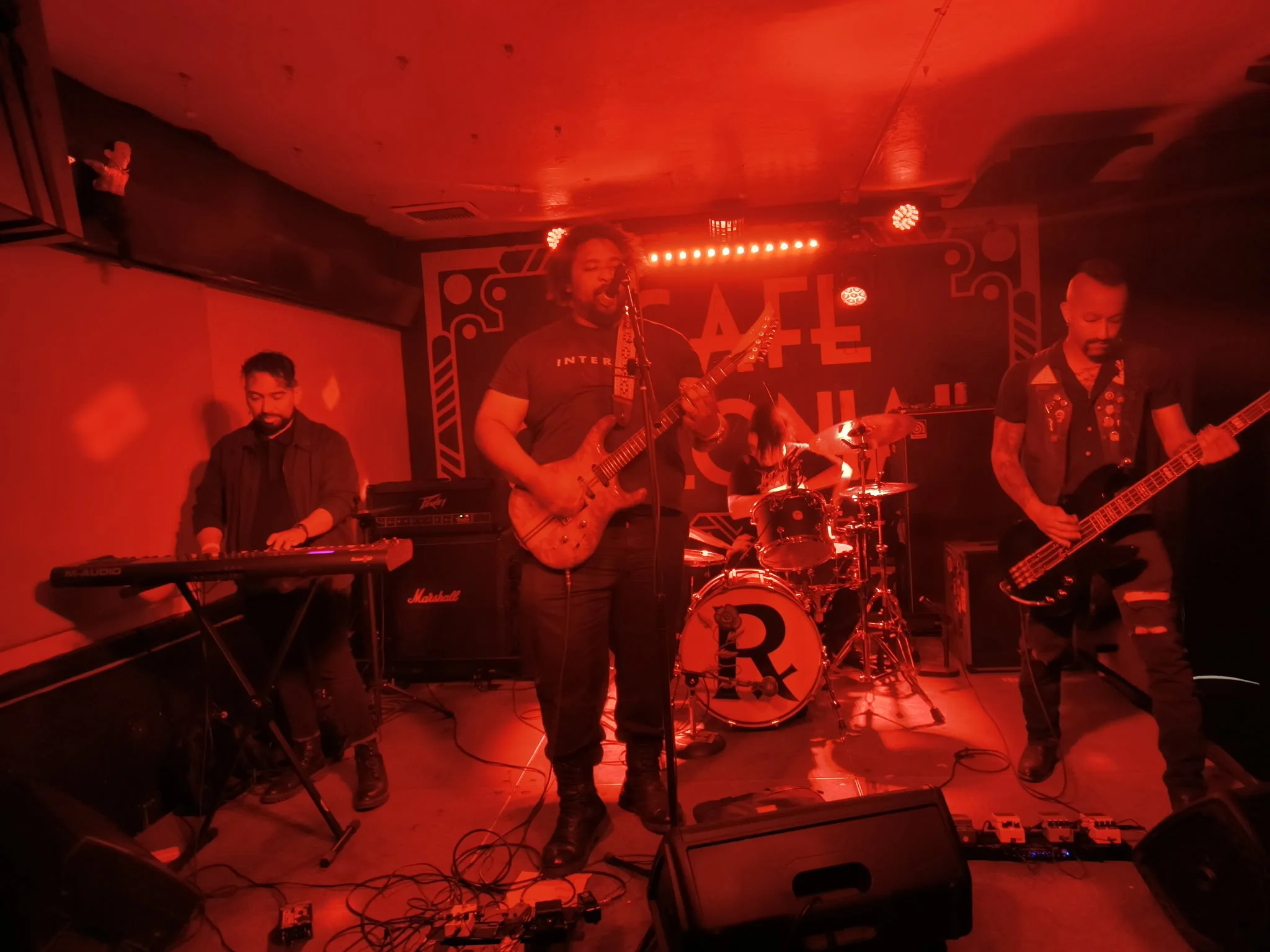 A band performing on stage with red lighting, featuring a guitarist singing into a microphone, a keyboardist, a bassist, and a drummer.