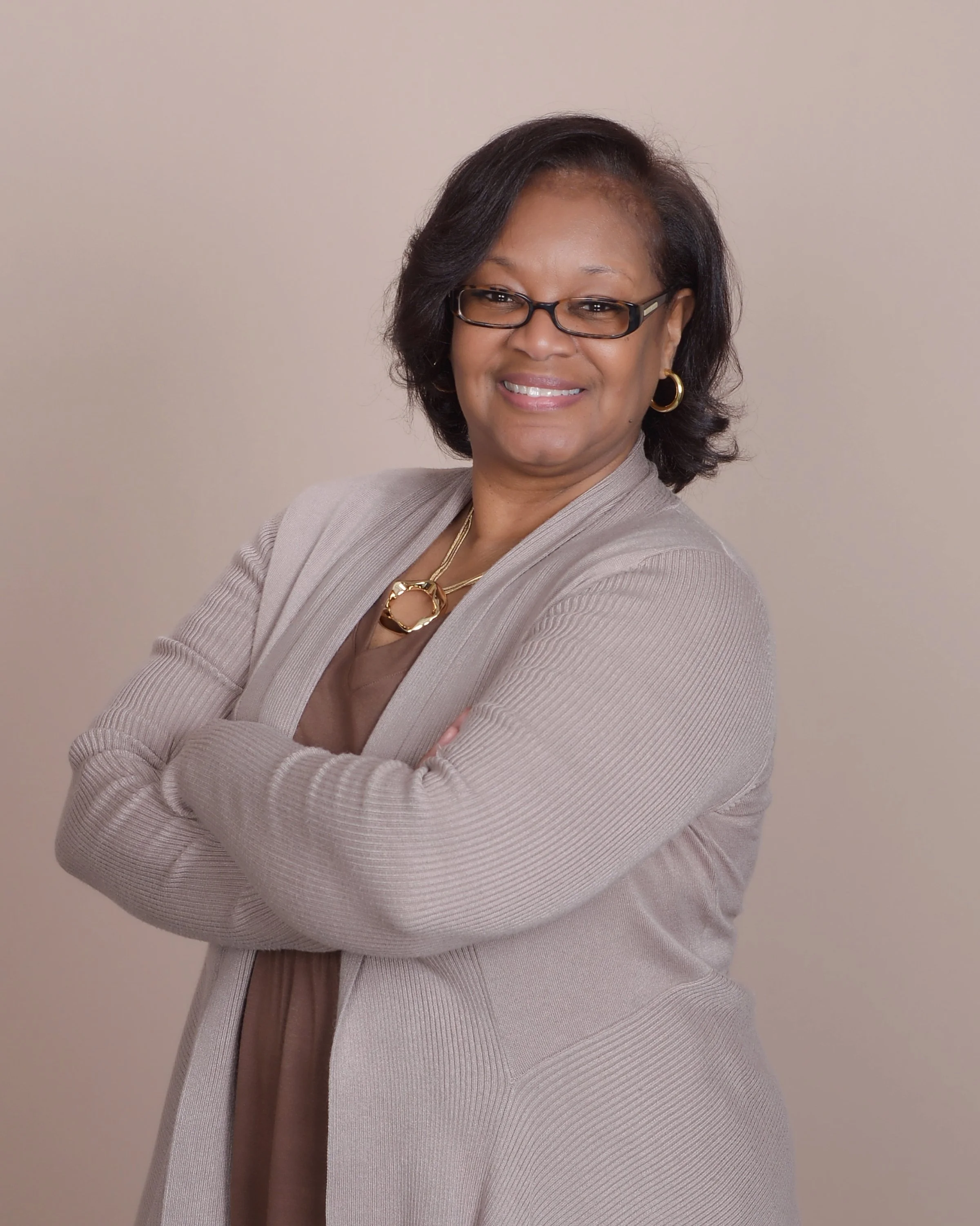 A smiling woman with glasses, wearing earrings, a gold necklace, and a beige blazer, standing against a plain light-colored background.