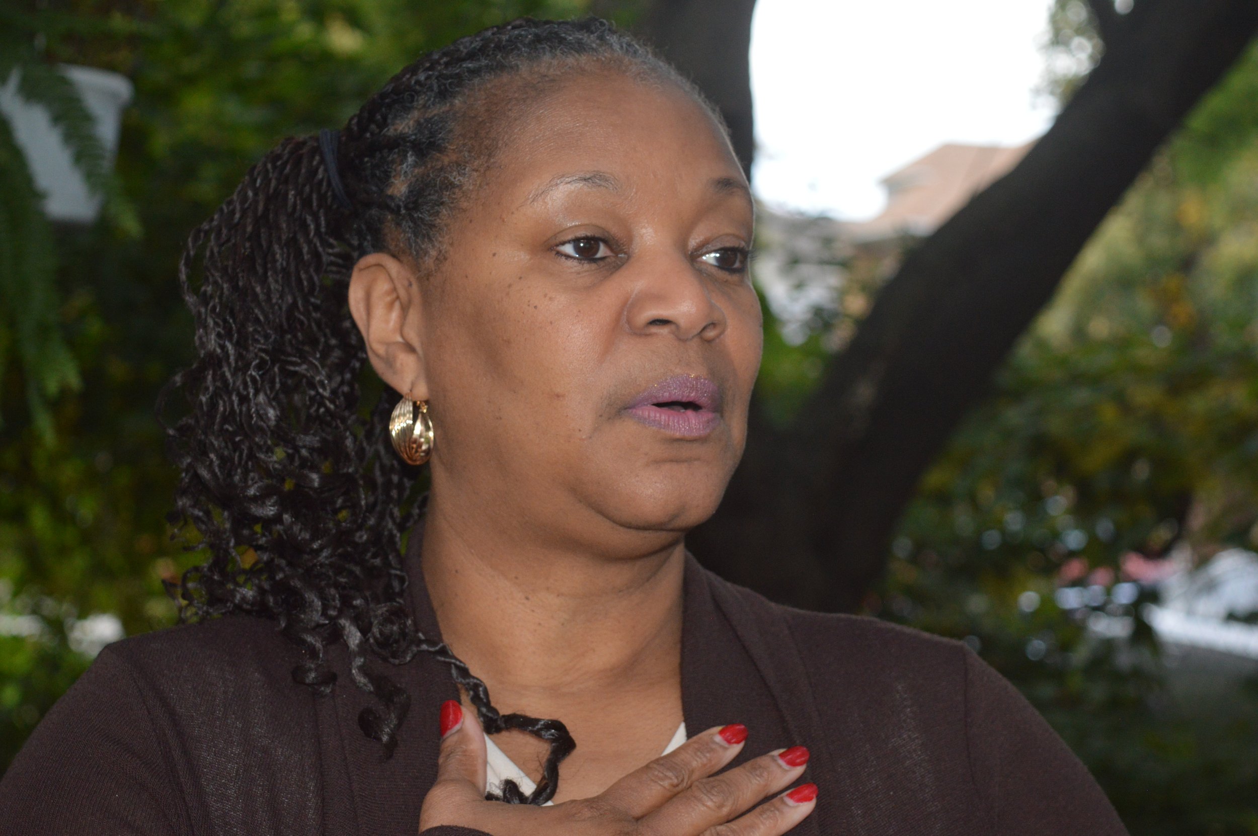 A woman with braided hair, wearing earrings and a dark blazer, has her hand over her chest during an outdoor event, with trees and houses in the background.