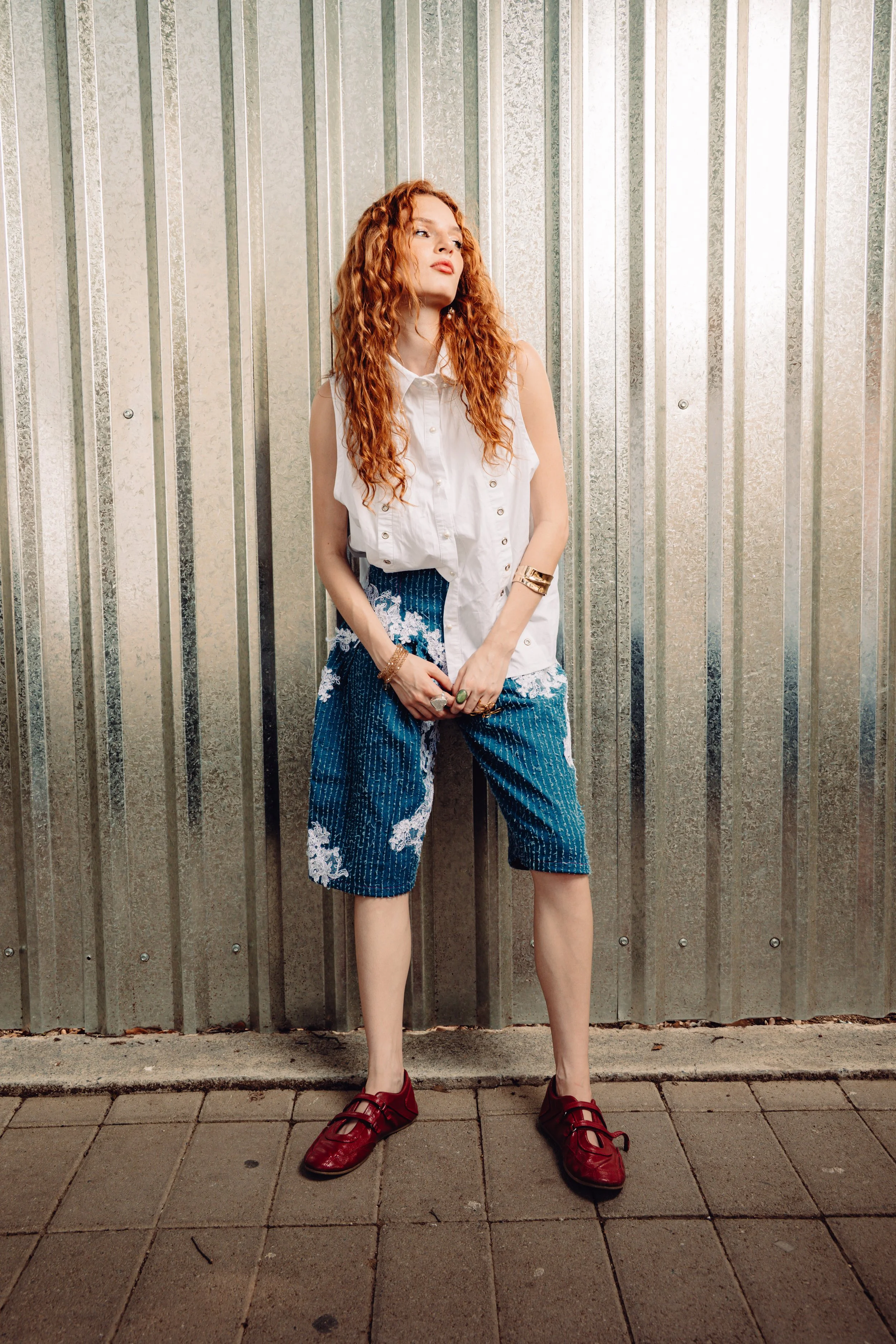 Modelo con blusa blanca, pantalón azul y zapatos rojos junto a un muro metálico.