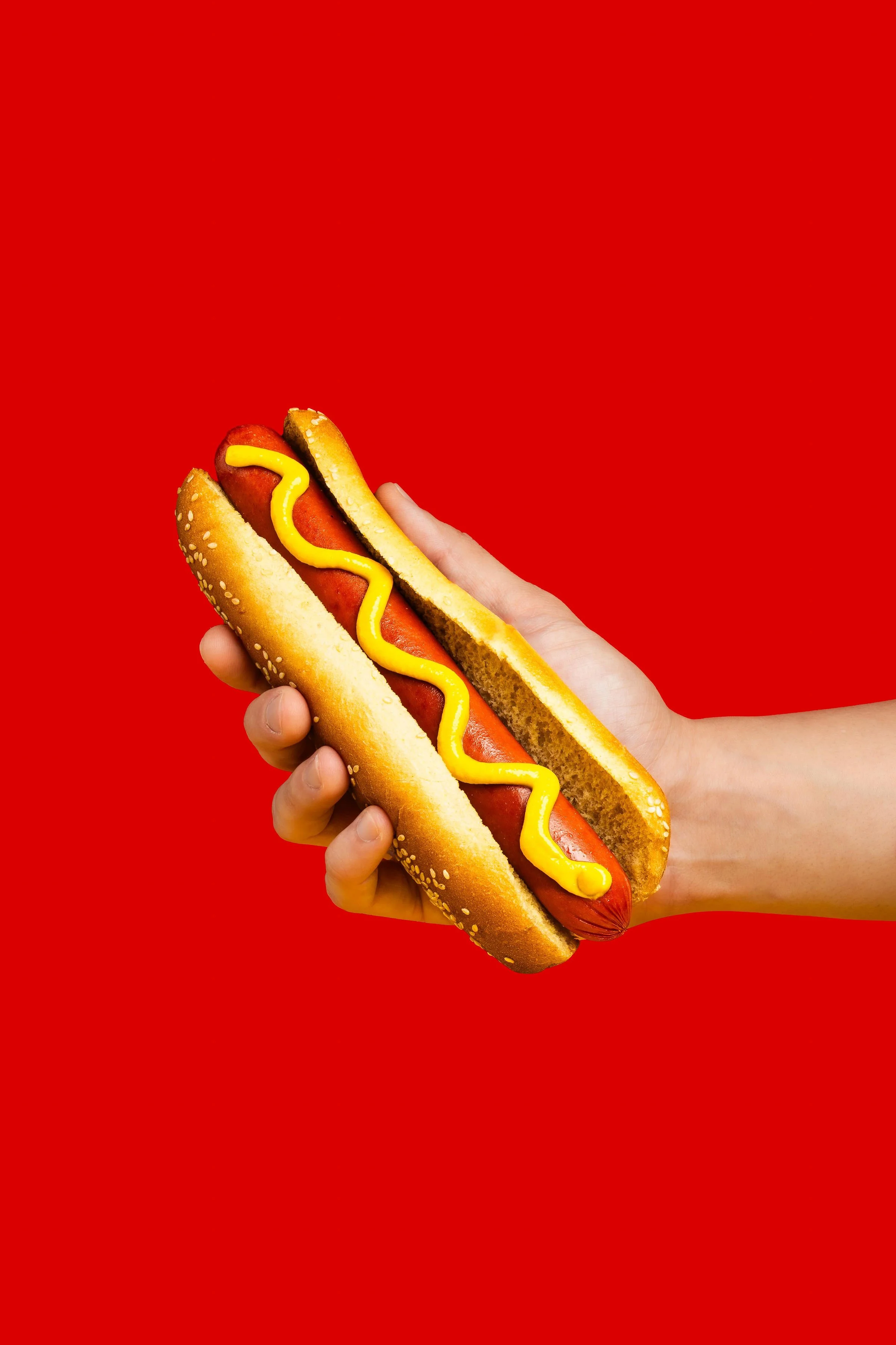 Mano sosteniendo un hot dog con mostaza sobre fondo rojo.