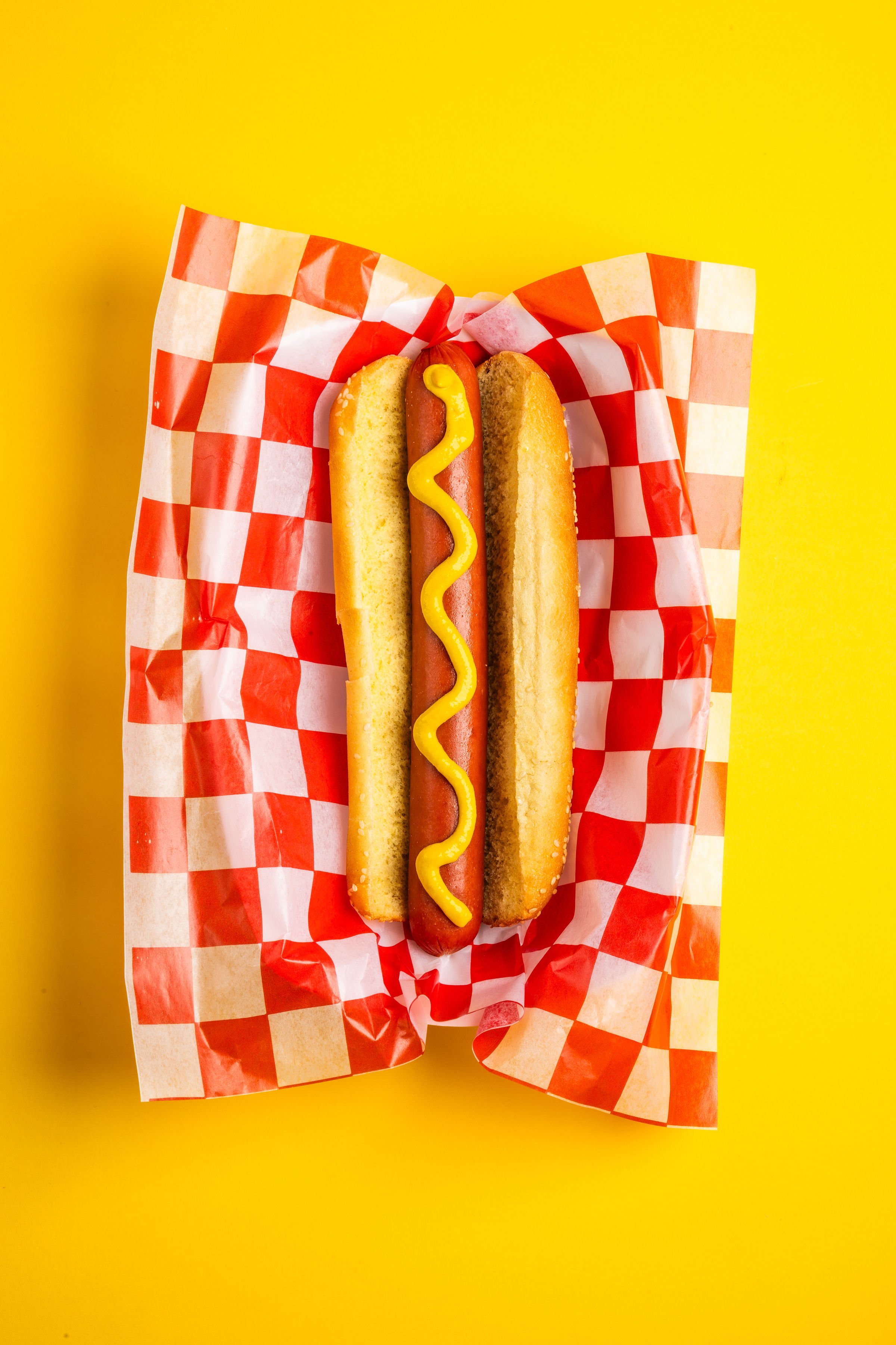 Hot dog con mostaza sobre papel a cuadros y fondo amarillo.