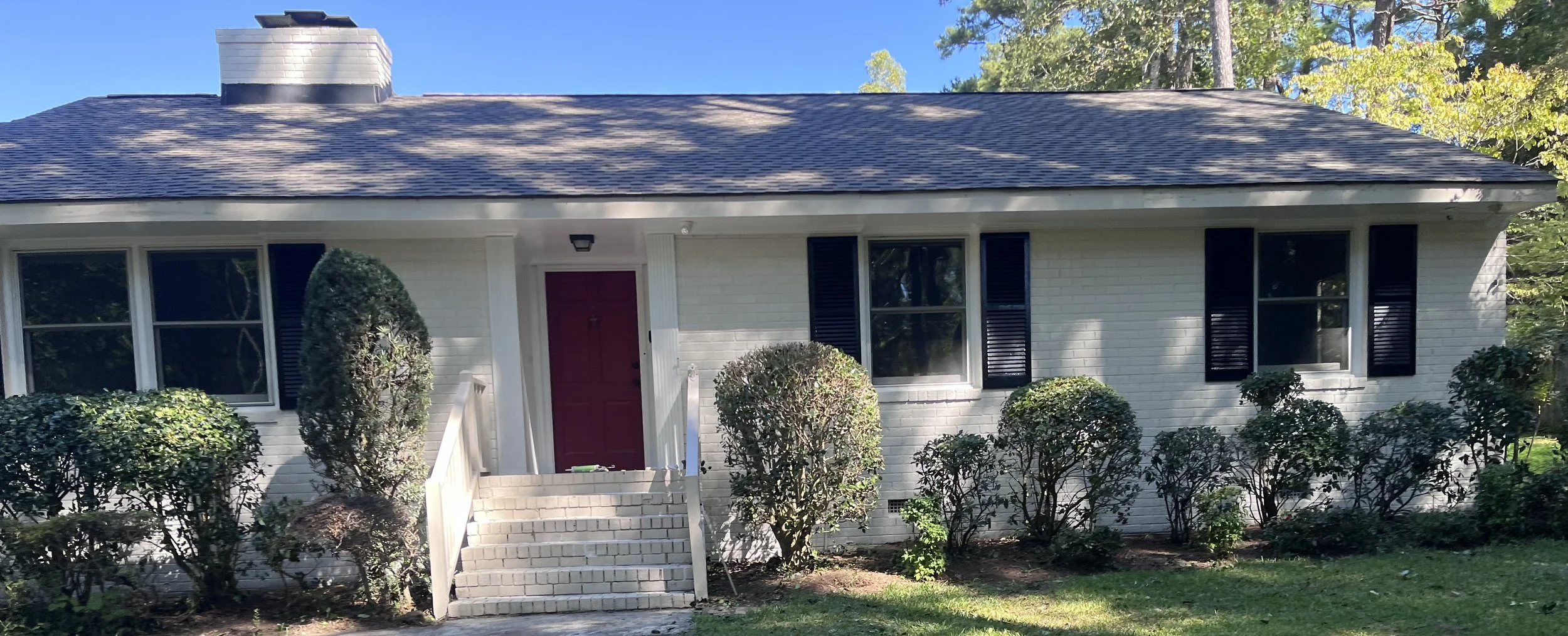 Front exterior view of a white brick house with a dark shingle roof and black window shutters, a red front door, a small staircase with white railing, and landscaped bushes in the front yard, under a clear blue sky.