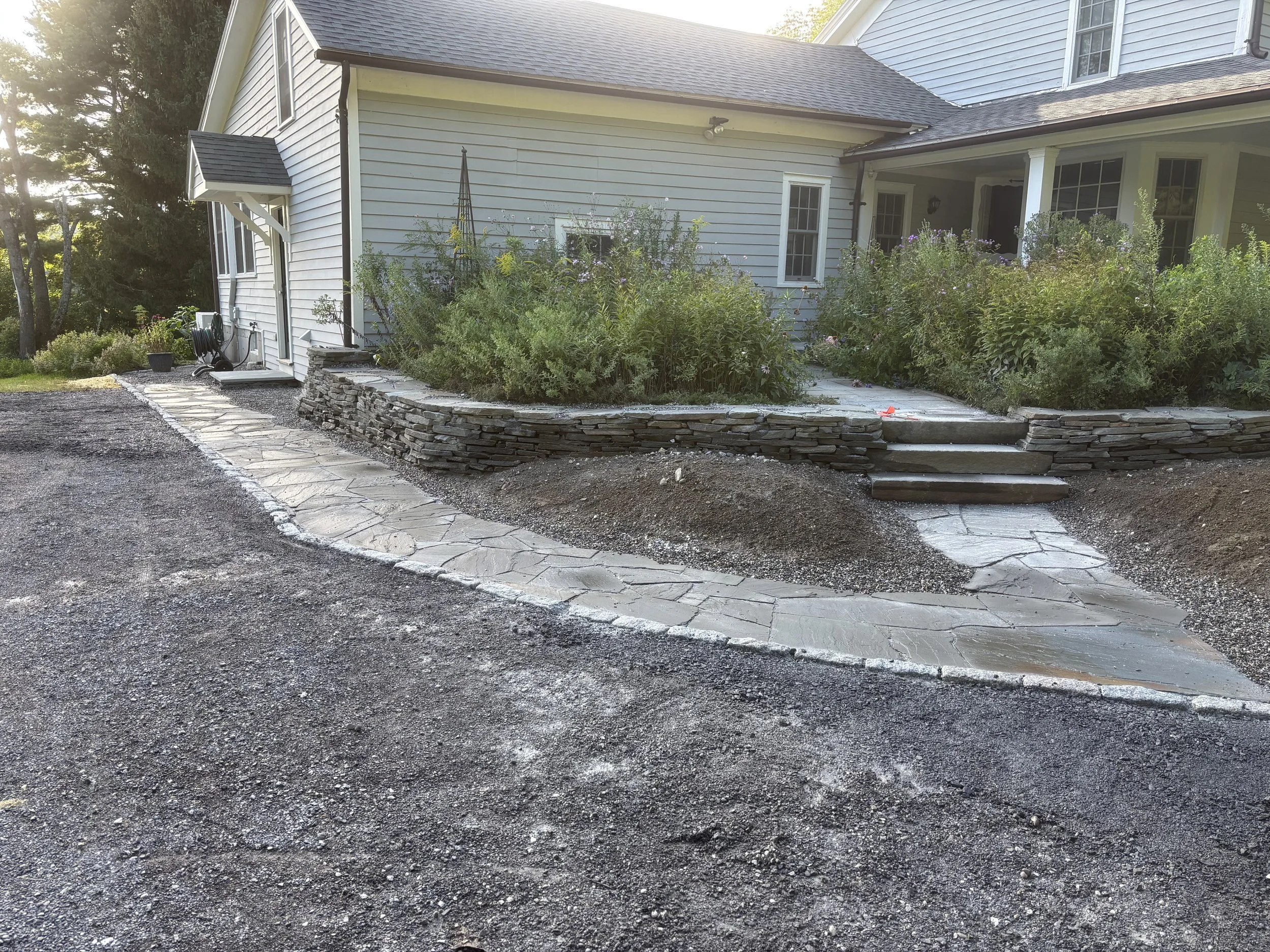The image shows the side yard of a house with a gravel driveway and a stone pathway. There are bushes and plants along the side of the house, which has white siding. A garden spray reel is on the wall, and a small set of concrete steps leads to a back porch. There is a large tree and sunlight in the background.