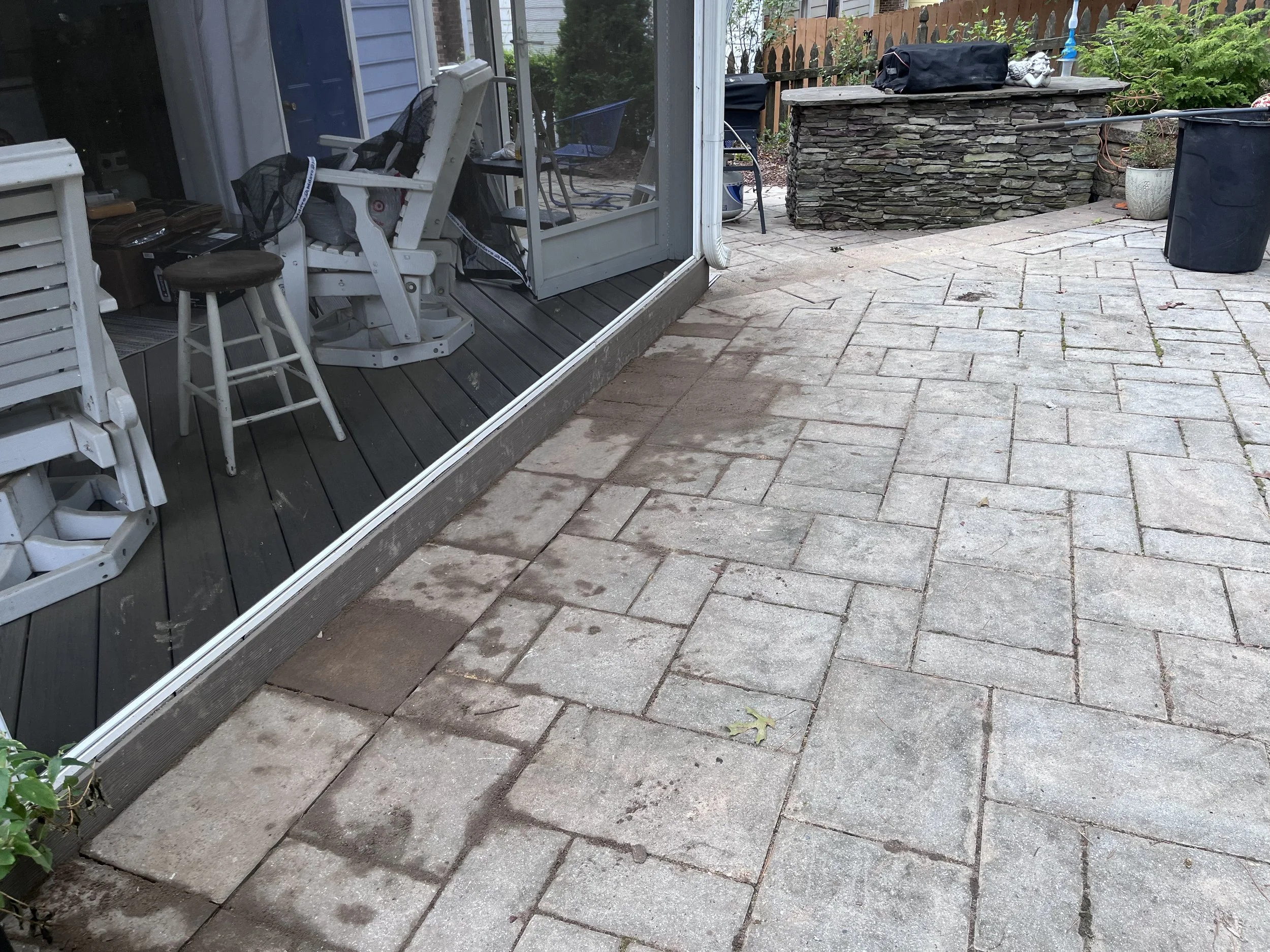 Backyard patio with stone pavers, outdoor furniture including a white Adirondack chair, and a stone feature wall, with gardening supplies and trash bins nearby.